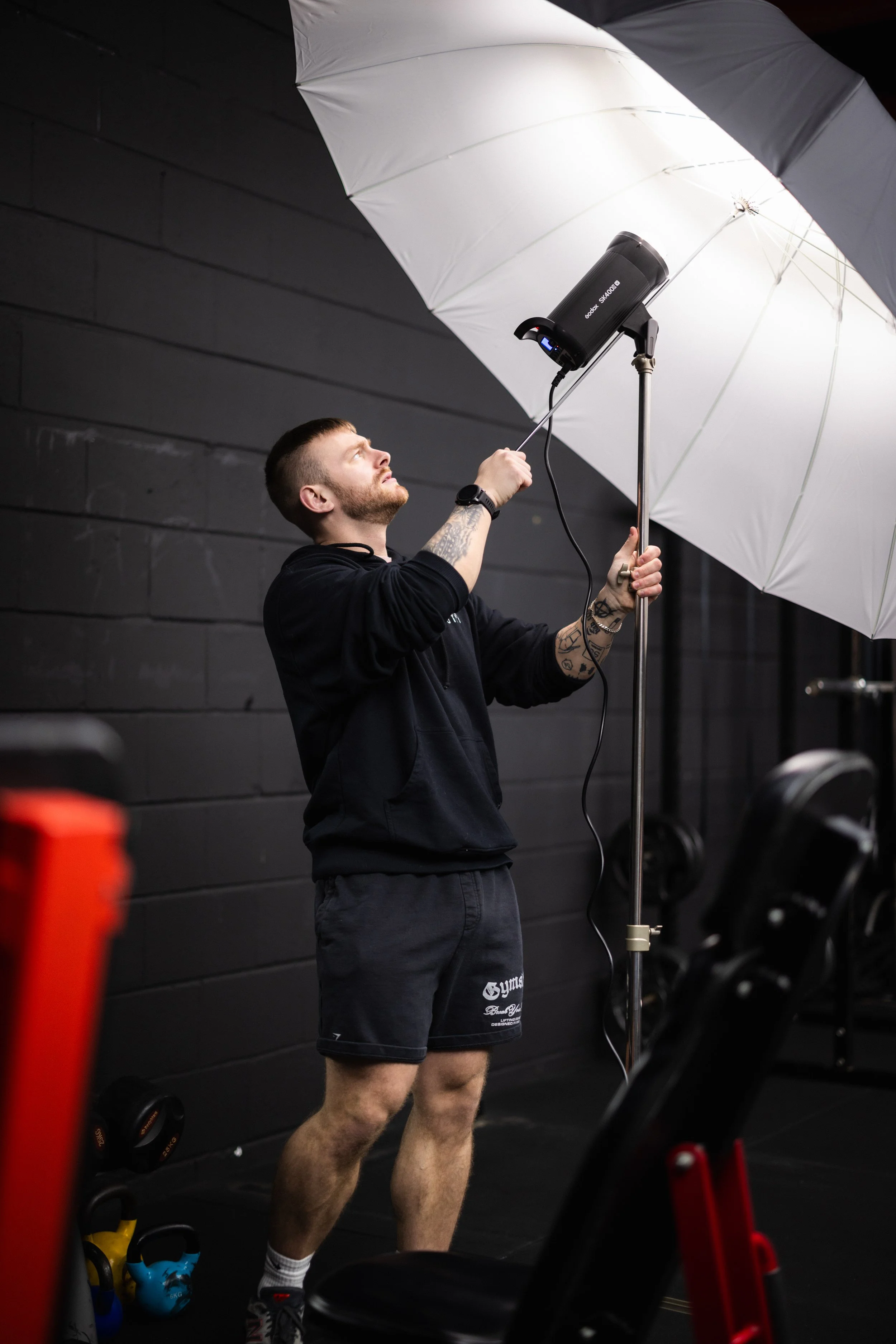 A man is adjusting a lighting setup with a white umbrella in a gym, indicating a photoshoot or video recording taking place.