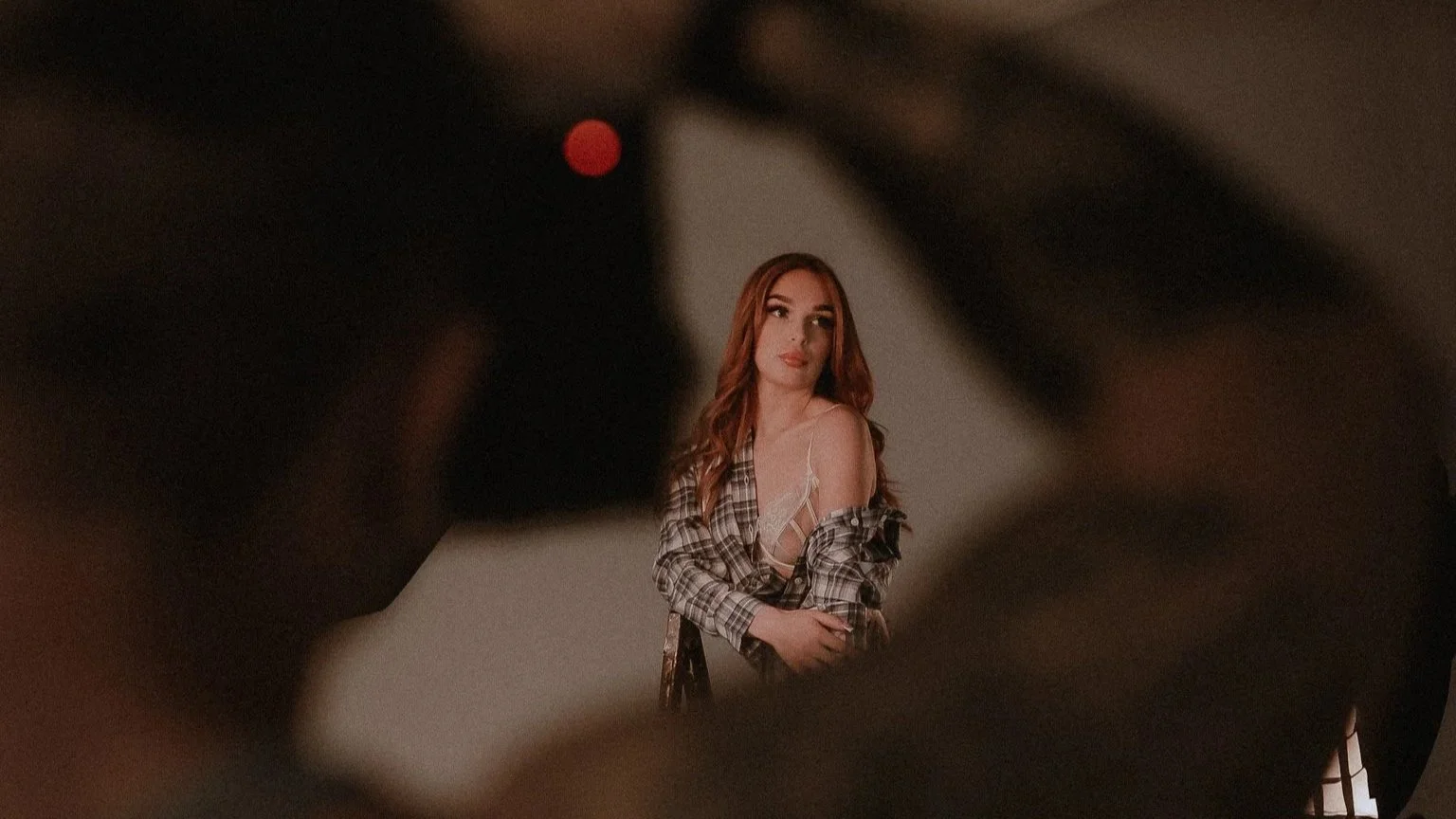 A woman with long red hair, wearing a plaid shirt and lace camisole, poses in front of a plain wall, viewed through an out-of-focus object.