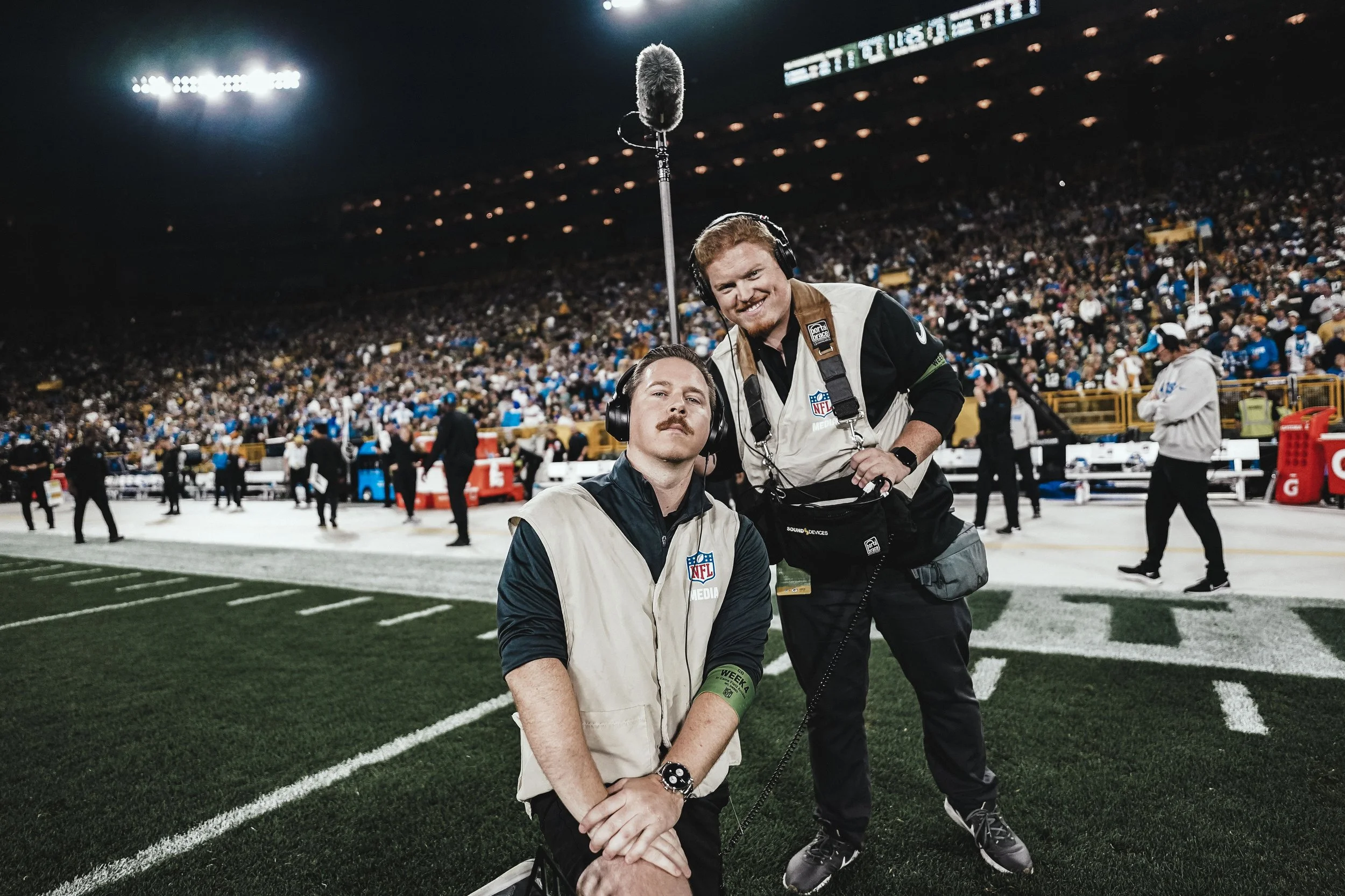 Two camera operators from NFL media on the sideline of a football field, with a large crowd in the stadium background, some wearing blue and white team apparel, under bright stadium lights.