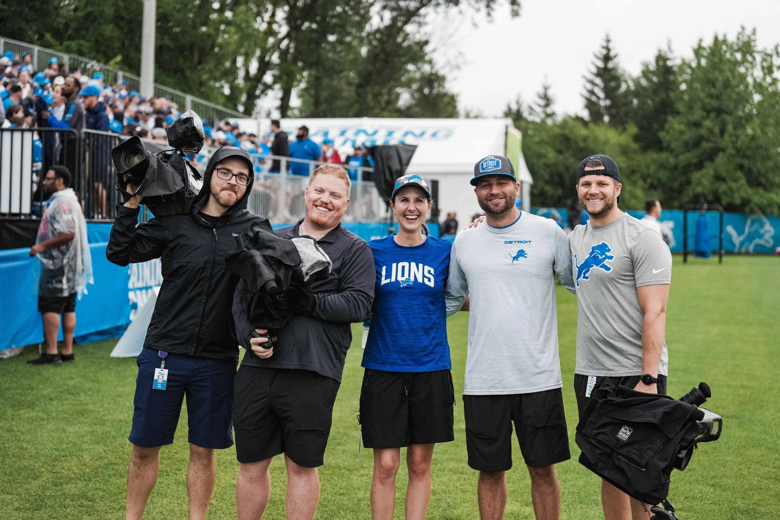 Five people standing outdoors on a sports field, smiling at the camera, with spectators in the background. They are dressed in Detroit Lions team apparel, holding cameras and equipment.