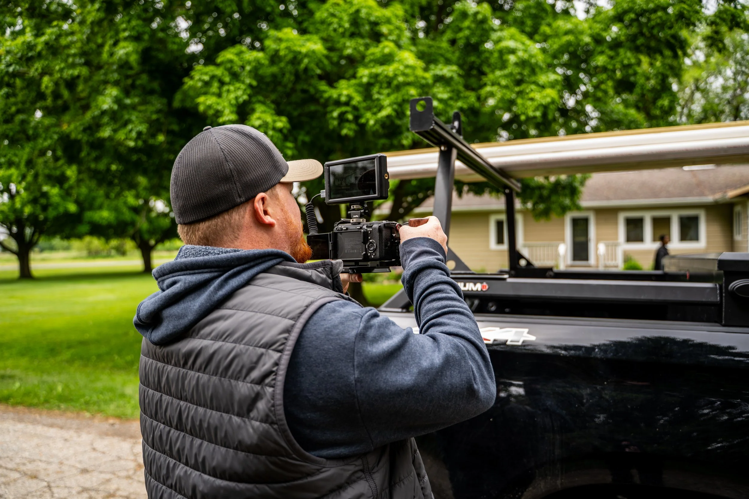 A man operating a camera mounted on a vehicle in a residential yard with green trees and a house in the background.