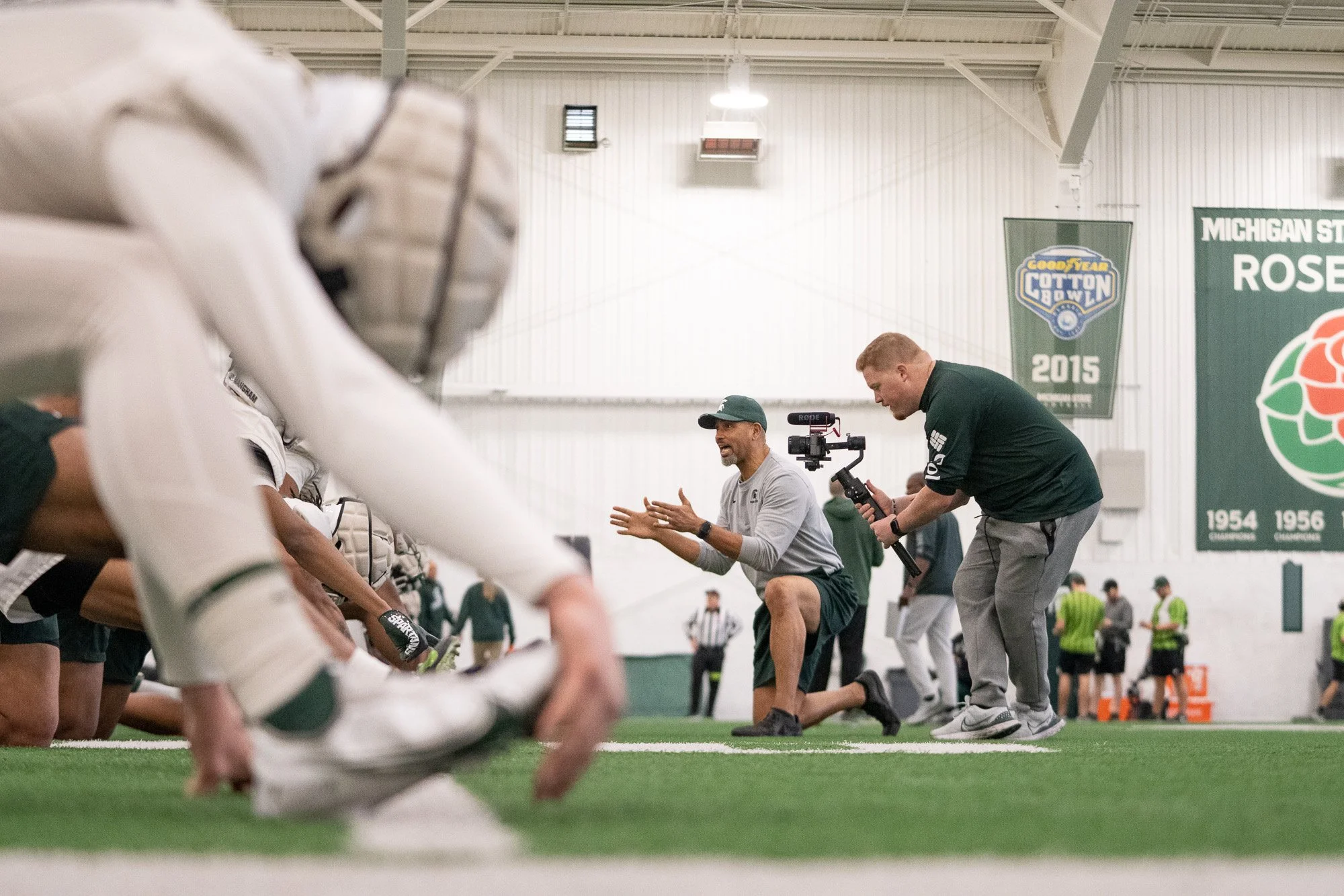Football players in a huddle during a practice or game at an indoor facility, with a coach kneeling and speaking to them, while a videographer records the scene.