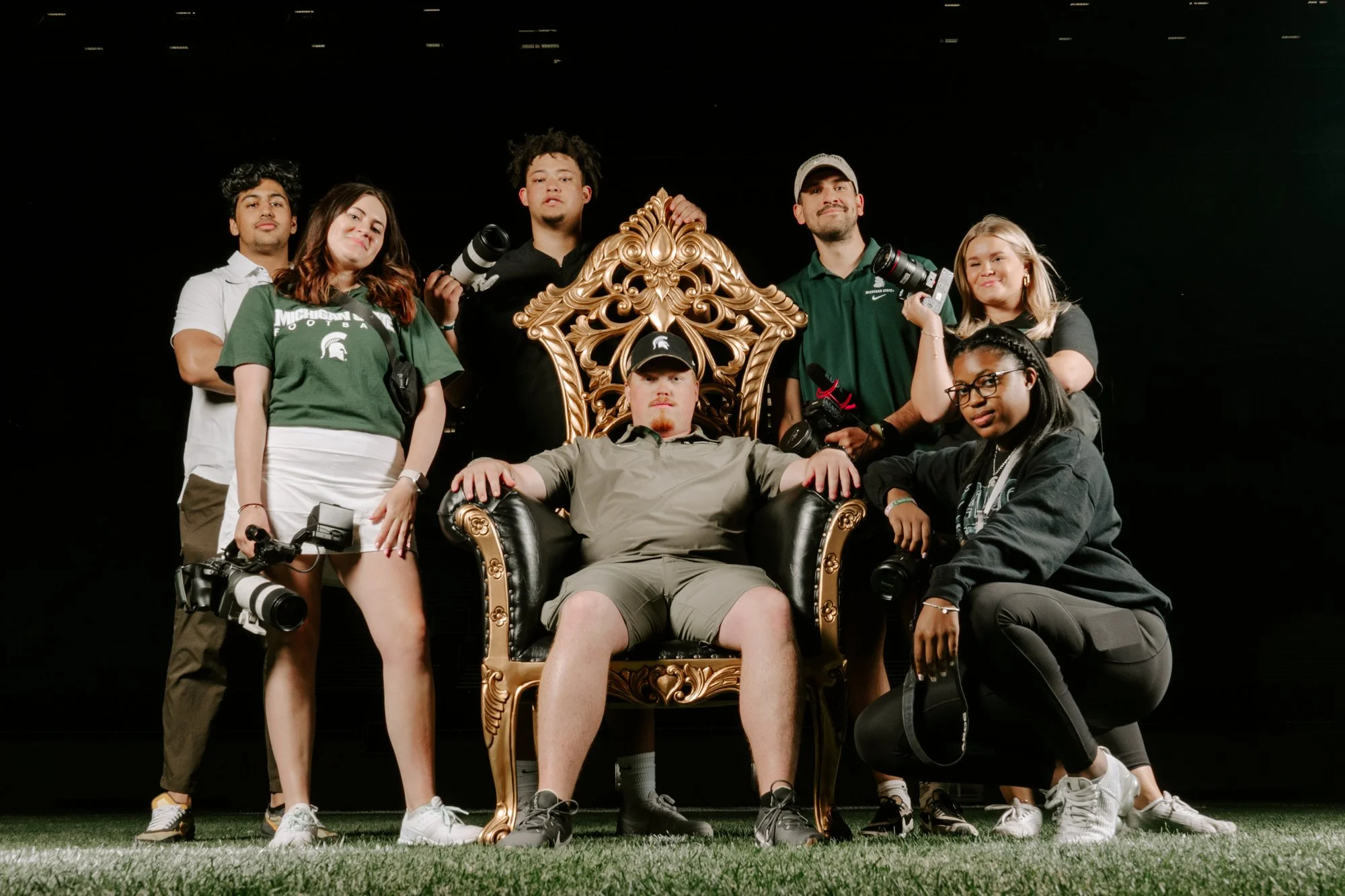 Group of young people, some holding cameras and lenses, gathered around a man sitting on an ornate, gold-trimmed black throne, against a dark background.