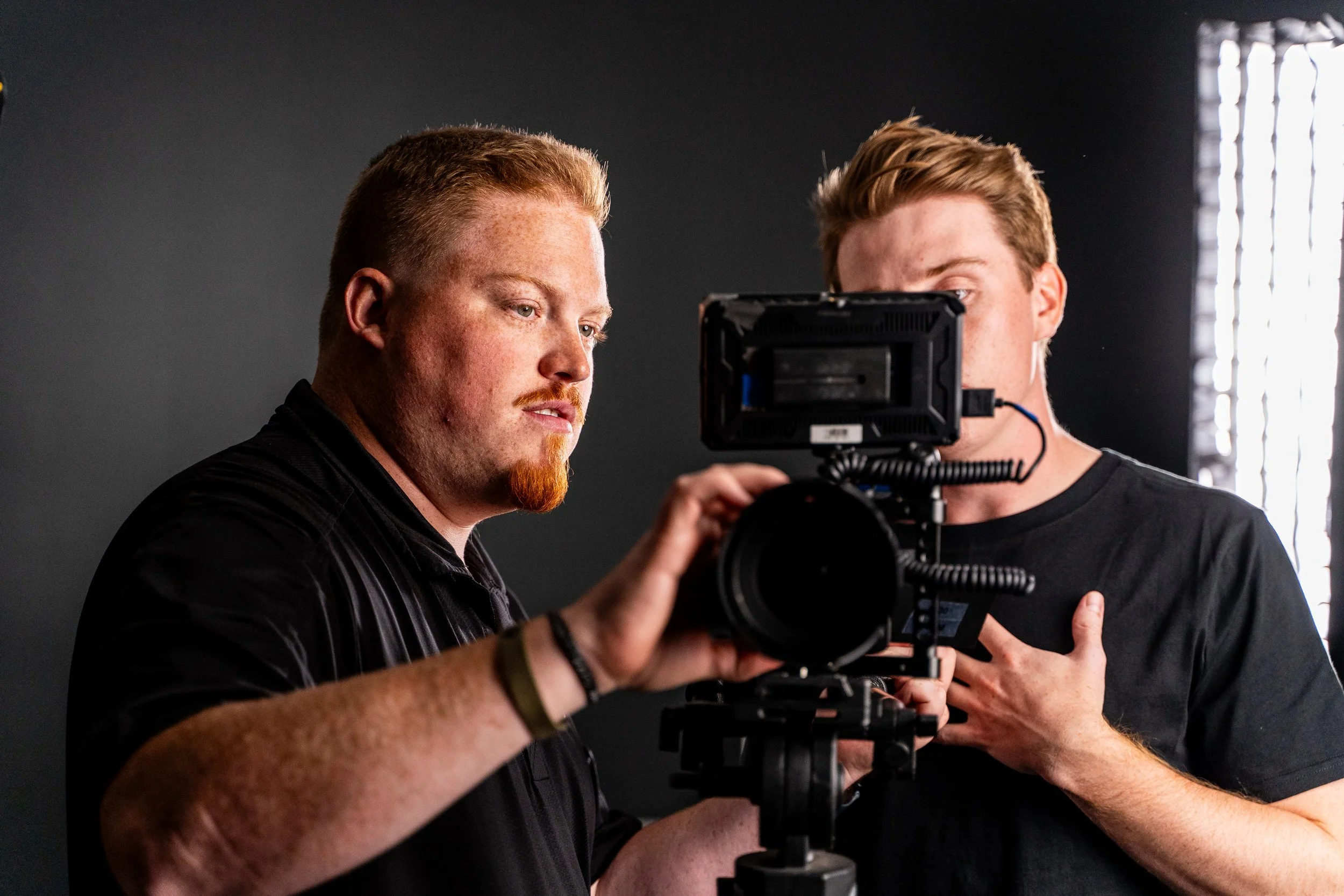 Two men operating a professional film camera in a studio with a dark background and bright lighting from the side.