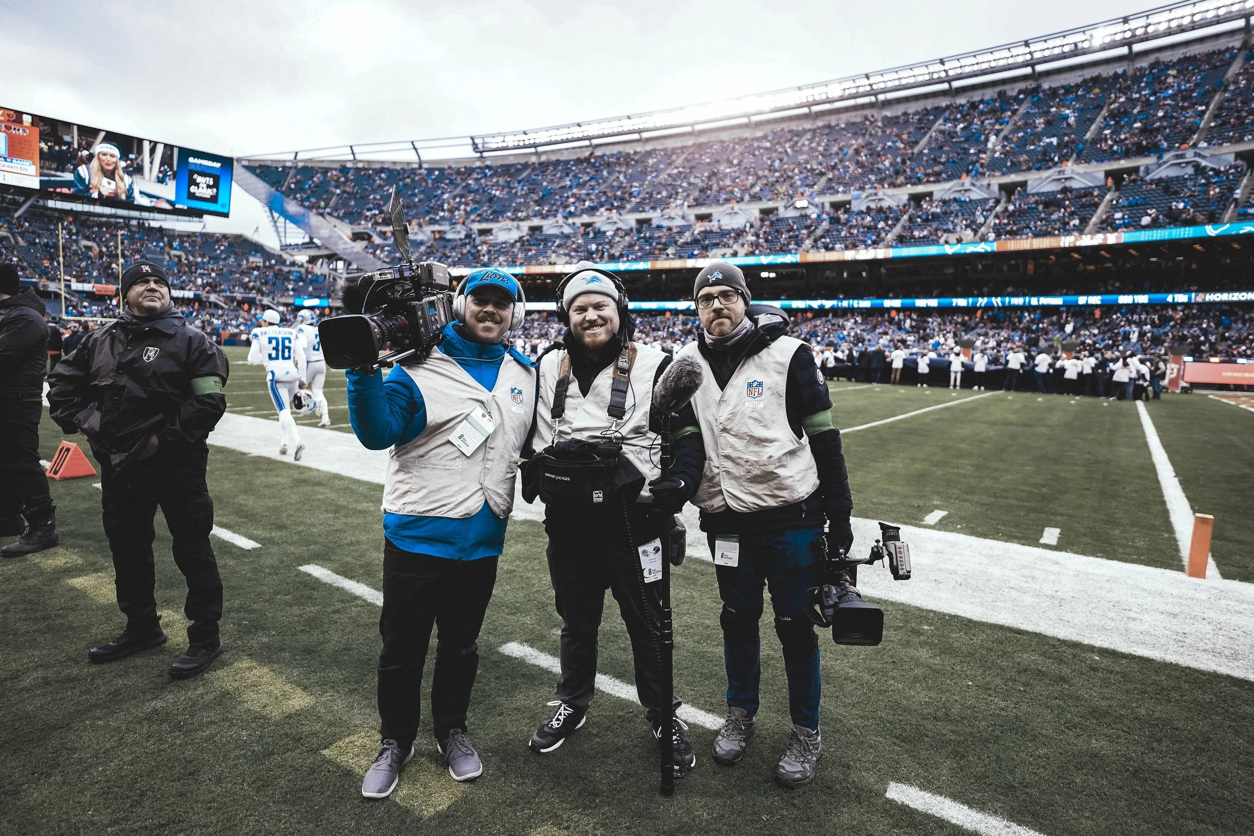 Three photographers standing on the sidelines of a football game field, smiling at the camera while holding cameras and equipment, with a stadium full of spectators in the background.