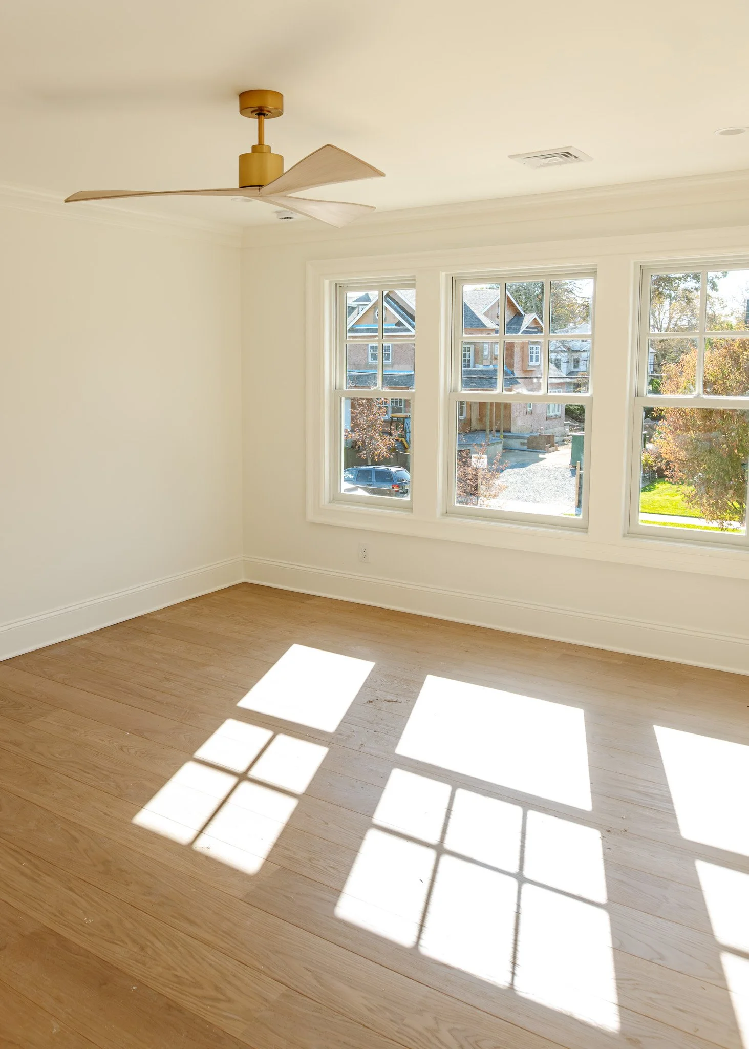 Habitación vacía con ventanas grandes que dejan entrar mucha luz natural, pisos de madera clara y un ventilador en el techo.