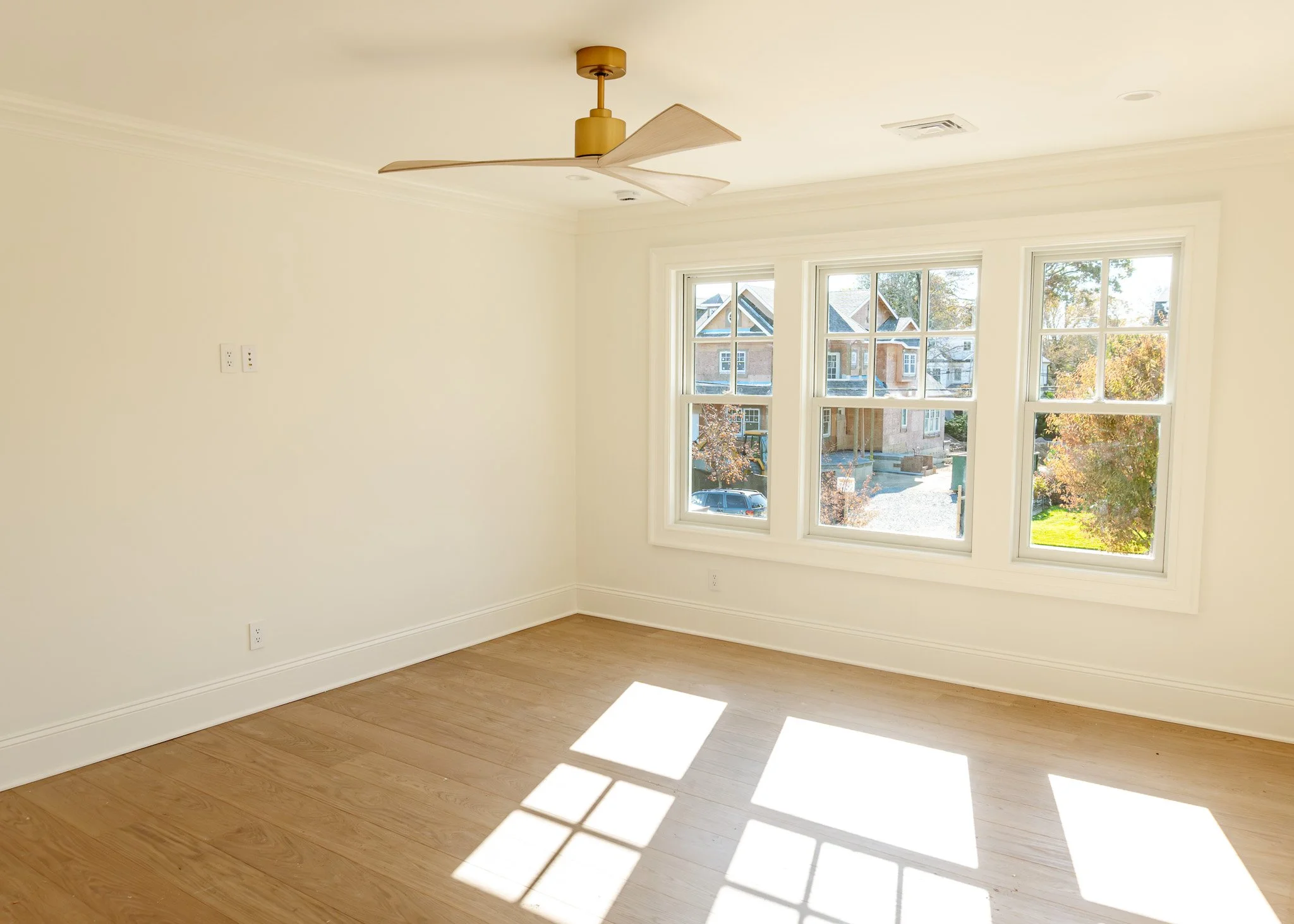 Habitación vacía con paredes blancas, suelo de madera y ventanas grandes que dejan entrar la luz solar, vista de casas vecinas y árboles afuera.