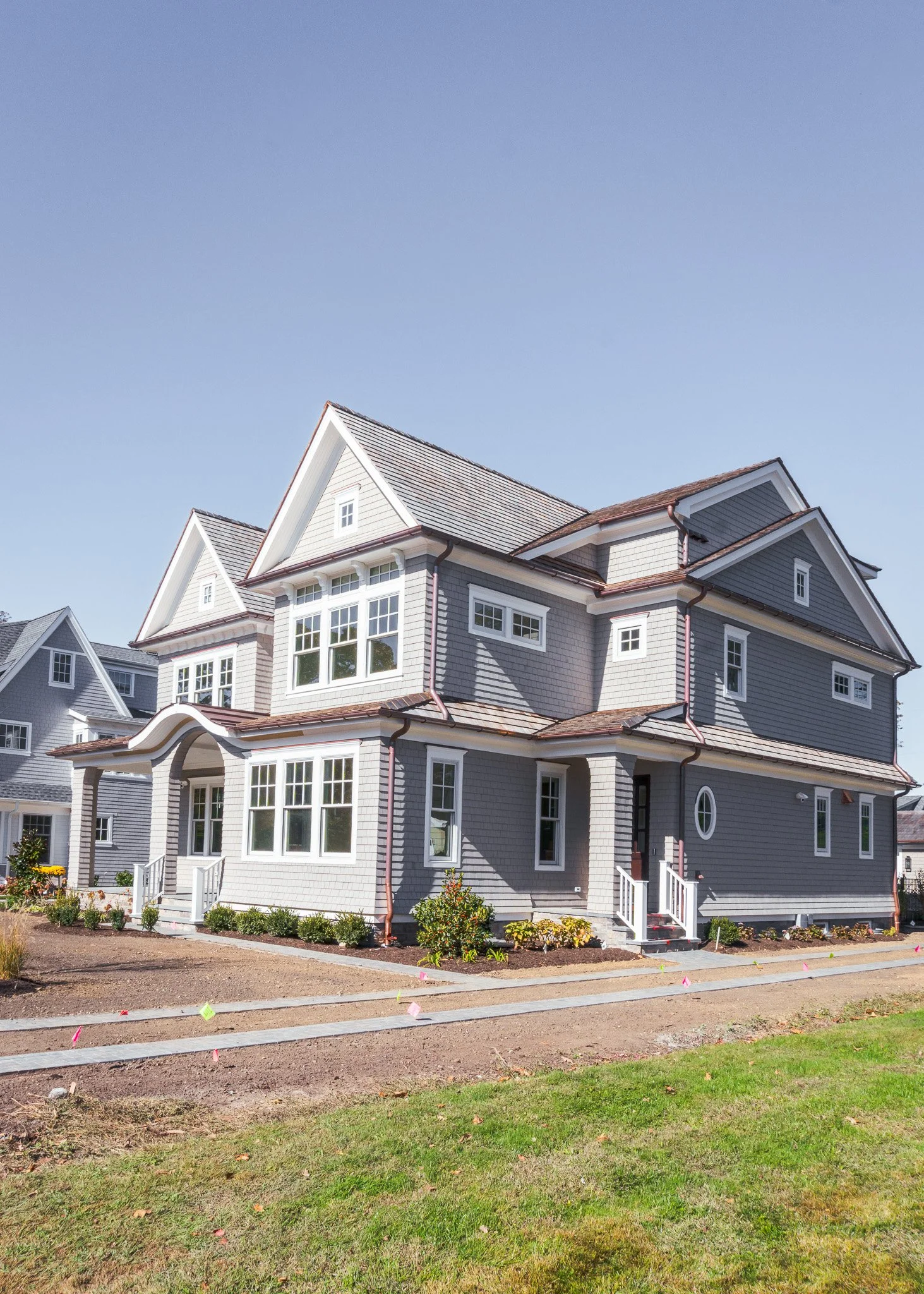Una casa de dos pisos de color gris con ventanas grandes y techos con tejas, en un espacio de césped y tierra con un camino de concreto y algunos arbustos y plantas alrededor.