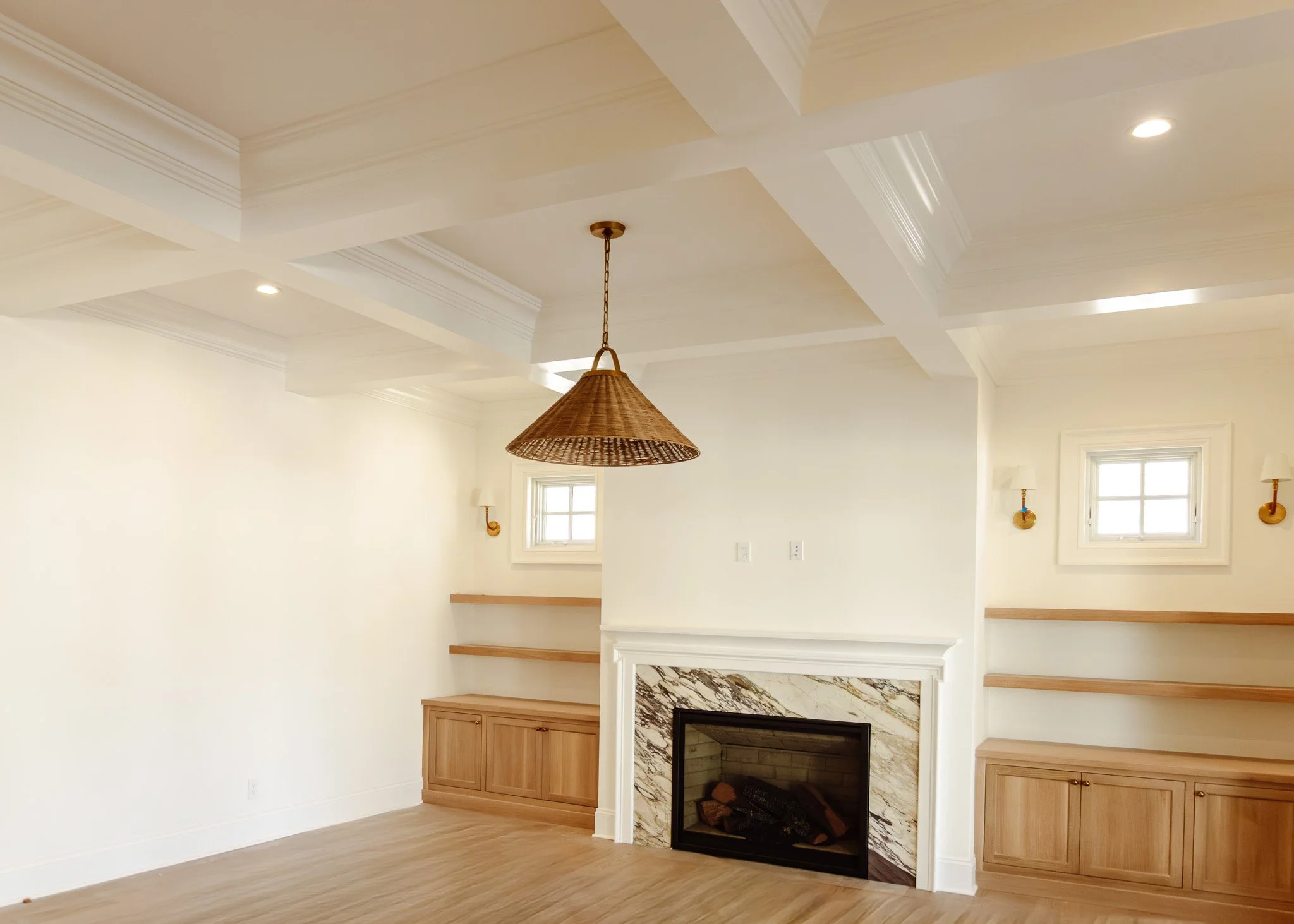 Sala con chimenea de mármol, paredes blancas, ventanas pequeñas y muebles de madera claro, lámpara de techo de mimbre y apliques de pared.