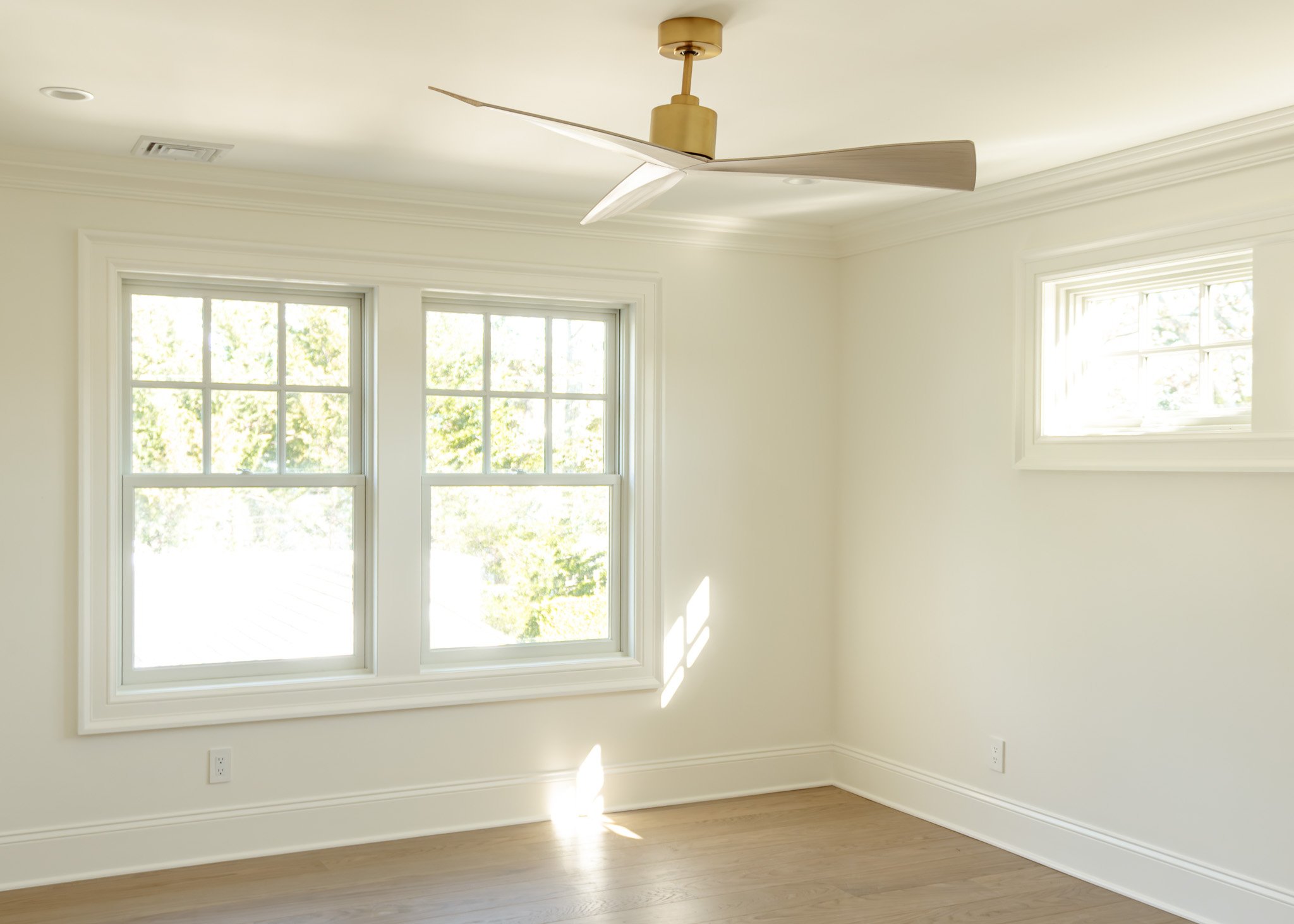 Habitación vacía con paredes blancas, dos ventanas grandes y una más pequeña, suelo de madera y ventilador en el techo.