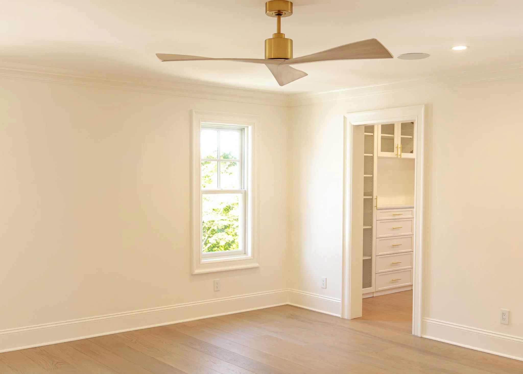 Habitación vacía con suelo de madera clara, paredes blancas, ventana con vista a árboles, sensor de luz en el techo, ventilador de techo dorado con aspas de madera.