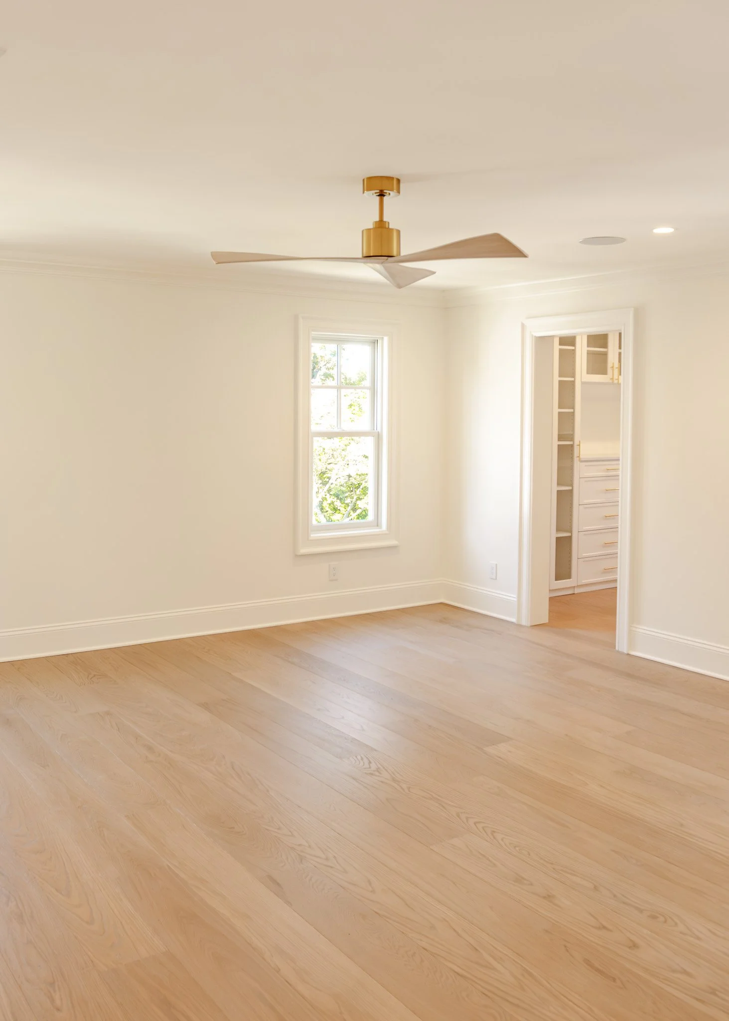 Habitación vacía con ventanas, techo con ventilador y suelo de madera clara.