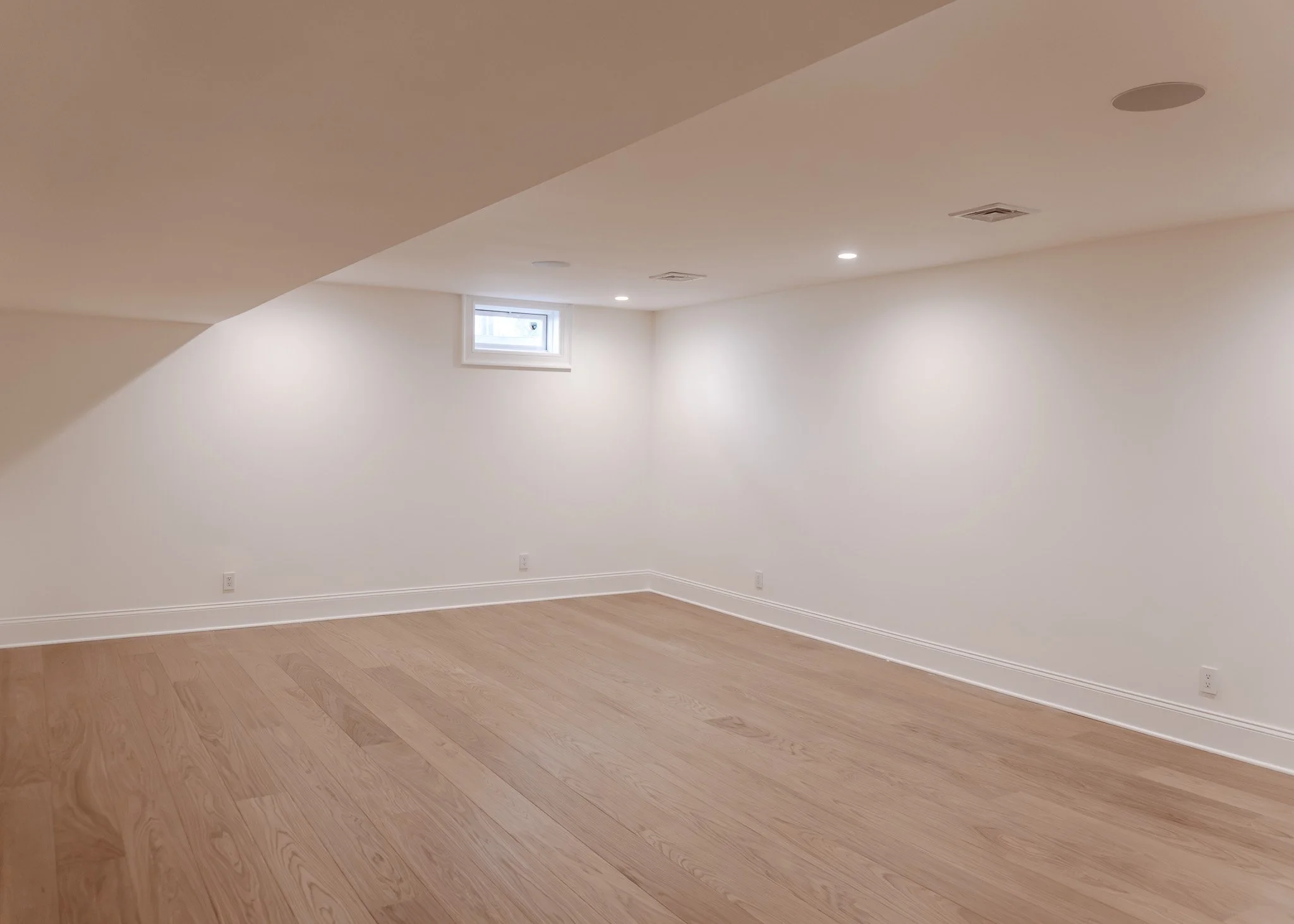 Espacio vacío con paredes blancas, suelo de madera y una pequeña ventana en la esquina superior, iluminación con luces empotradas en el techo.