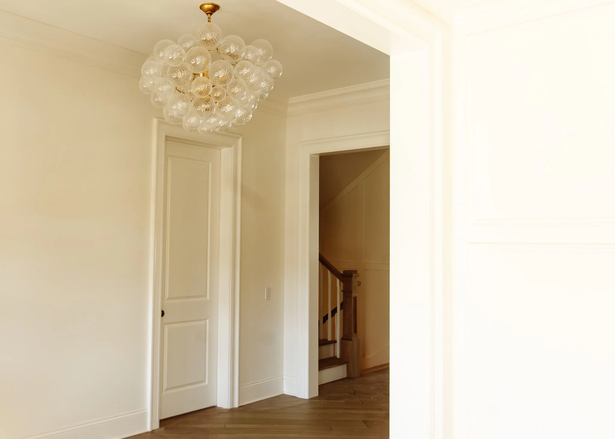 Interior de una casa con paredes blancas y un chandelier de bolitas de vidrio colgado del techo, una puerta blanca y una escalera de madera.