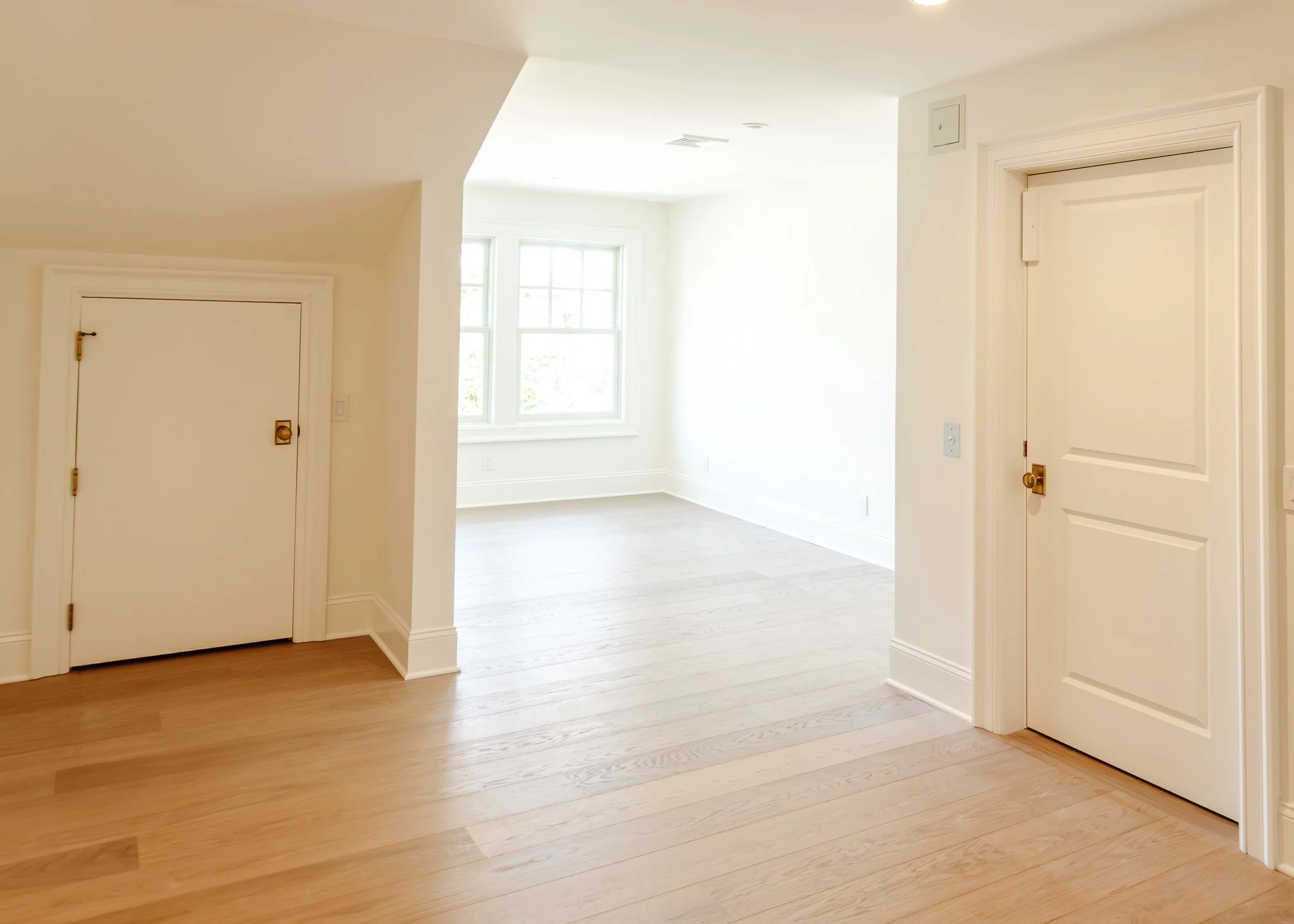Interior de una sala con paredes blancas, ventanas grandes que dejan entrar luz natural, piso de madera clara y puertas blancas con manijas doradas.
