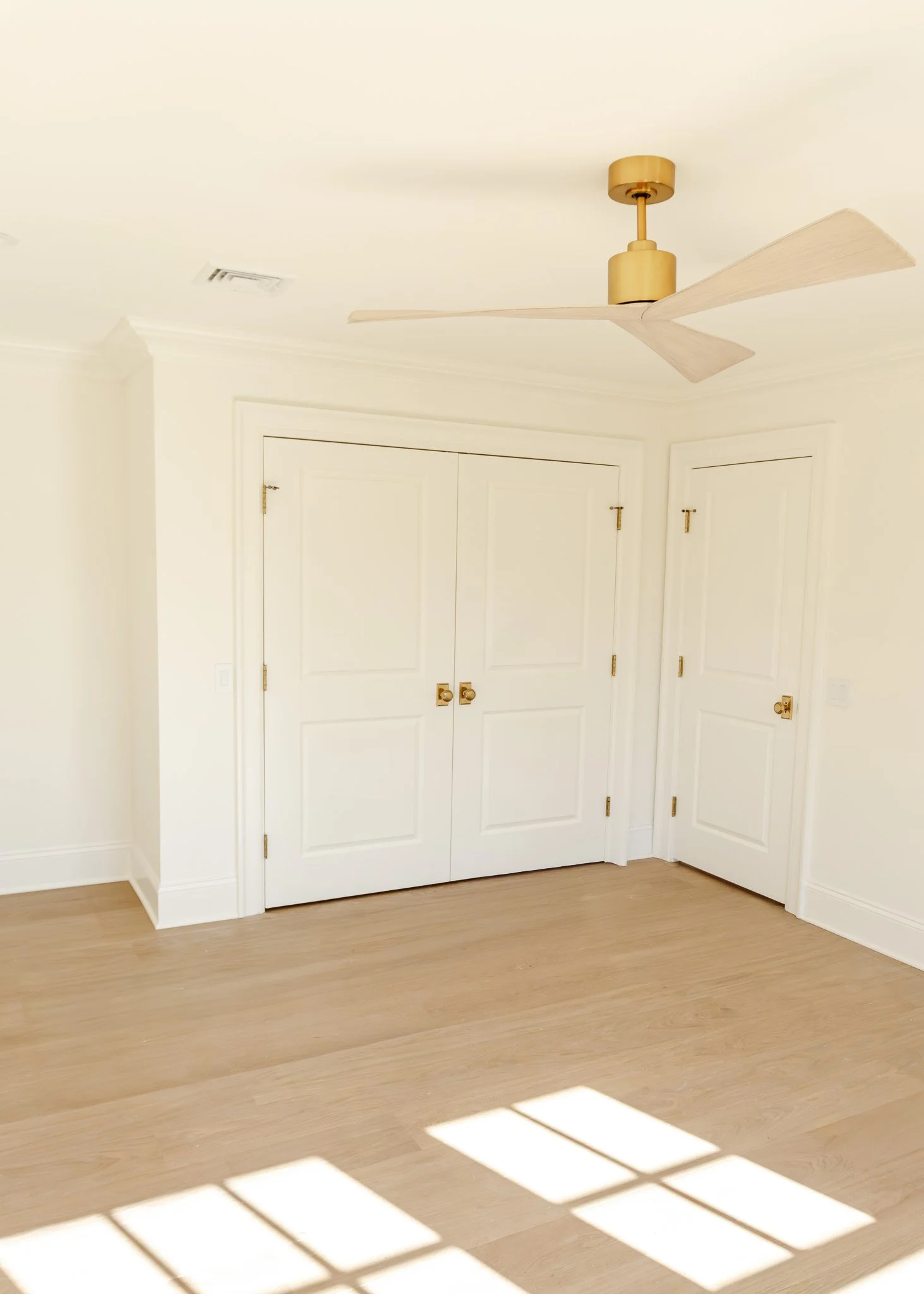 Habitación vacía con puertas blancas, suelo de madera clara y ventilador de techo dorado.