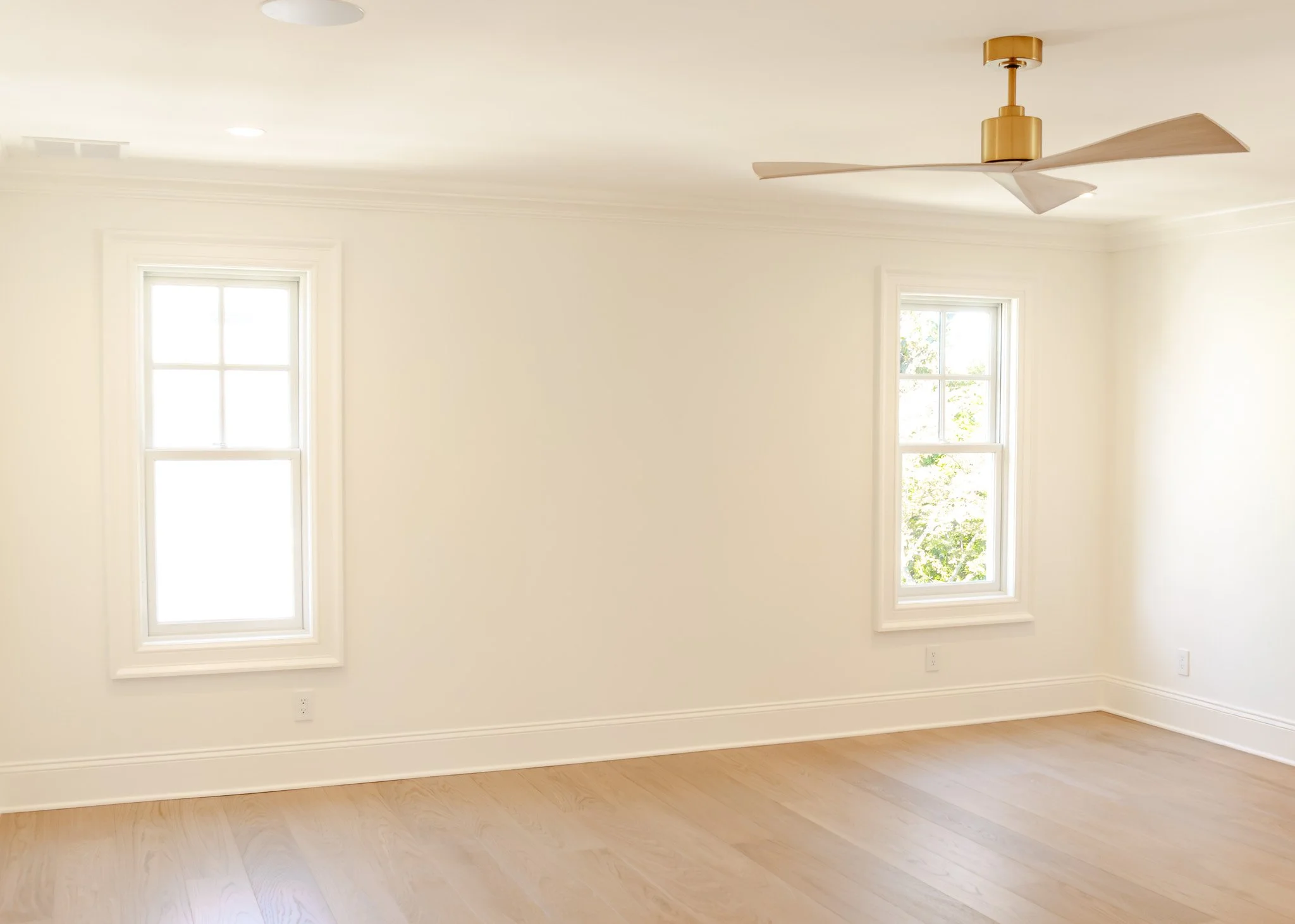 Habitación vacía con paredes blancas, dos ventanas, piso de madera y ventilador de techo dorado.