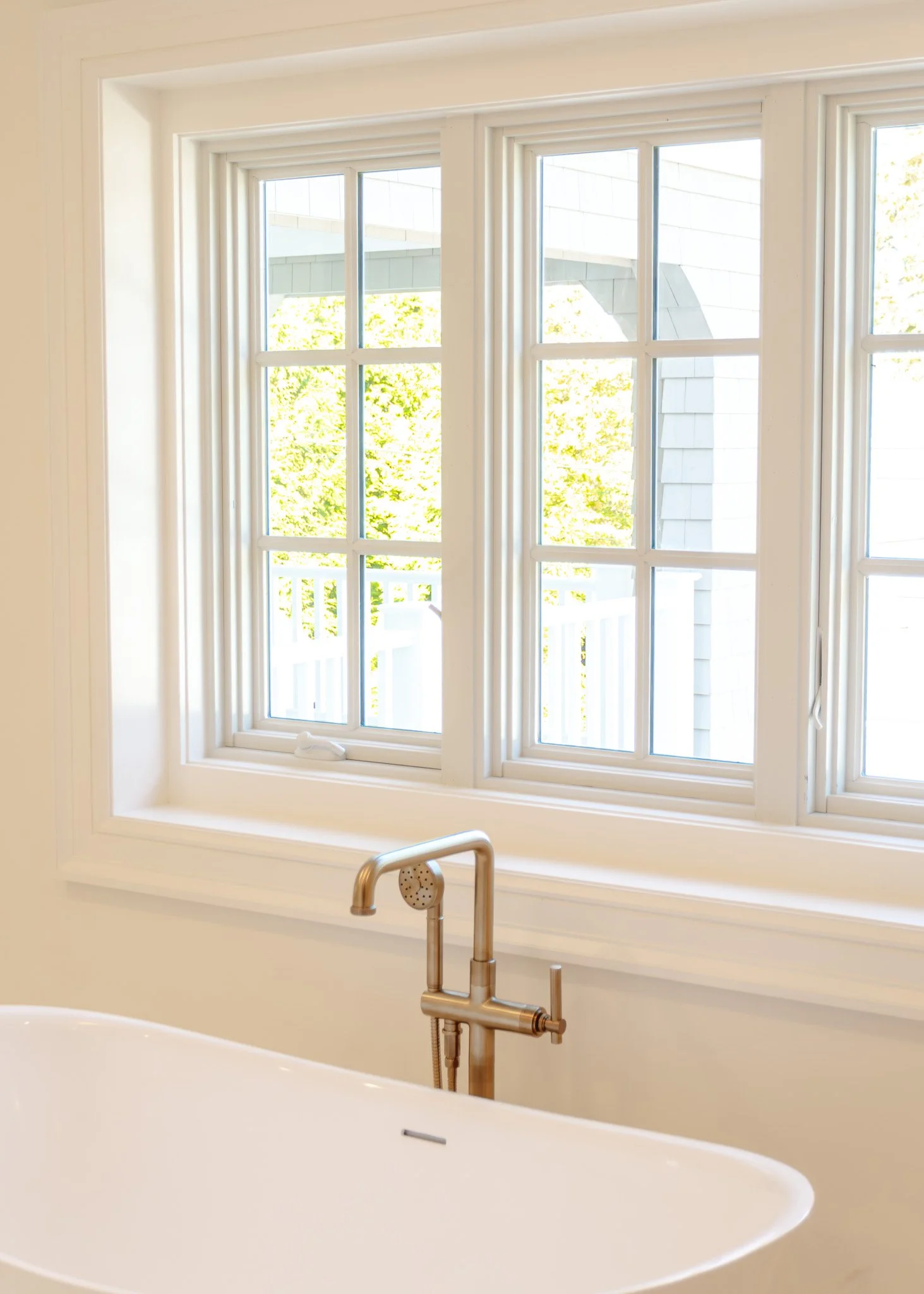Ventana grande con marco blanco y vista a un área con árboles verdes, y una bañera blanca con grifo de metal en un baño luminoso