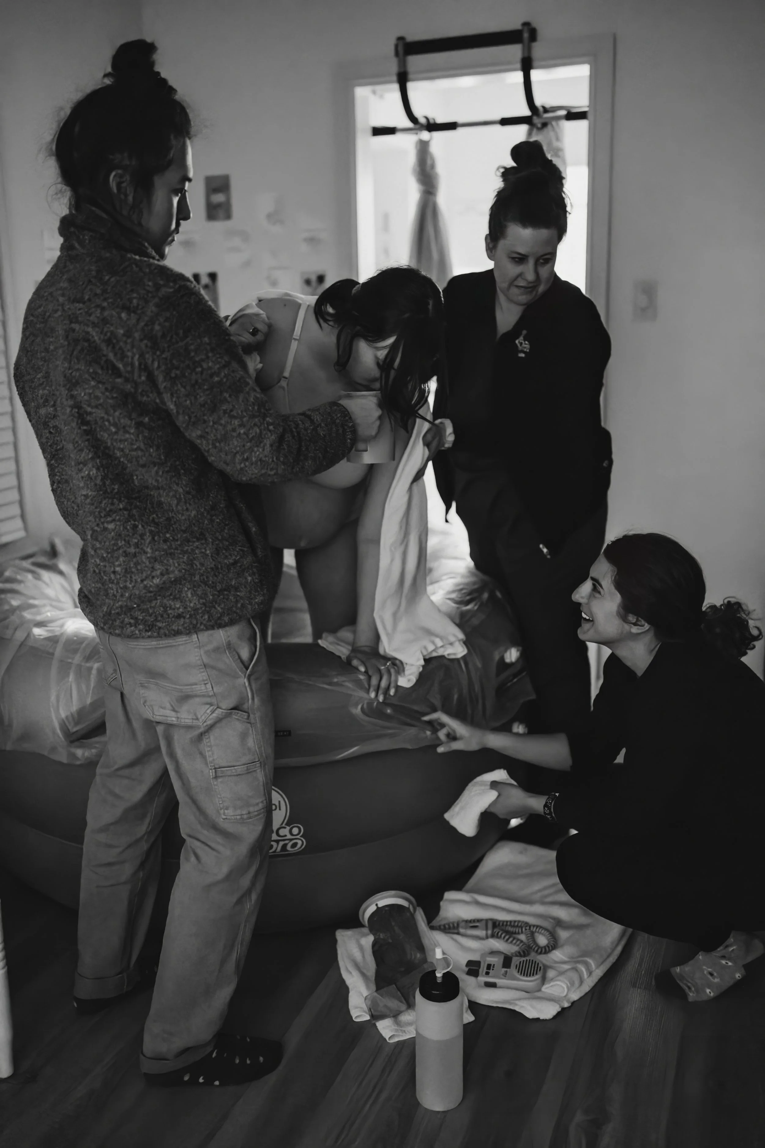 A group of four women attending a childbirth or midwifery class in a small room with a window, including one woman lying back in a birthing pool and a woman sitting nearby. The scene appears to be instructional or supportive, with the women engaging in a demonstration or practice.