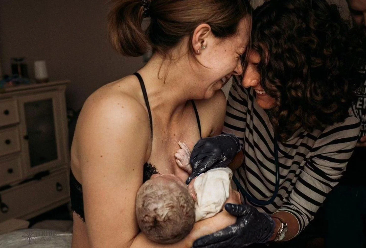 A woman has just given birth and holds her newborn baby while a woman in striped clothing supports her; birthing environment appears to be in a home or hospital setting.