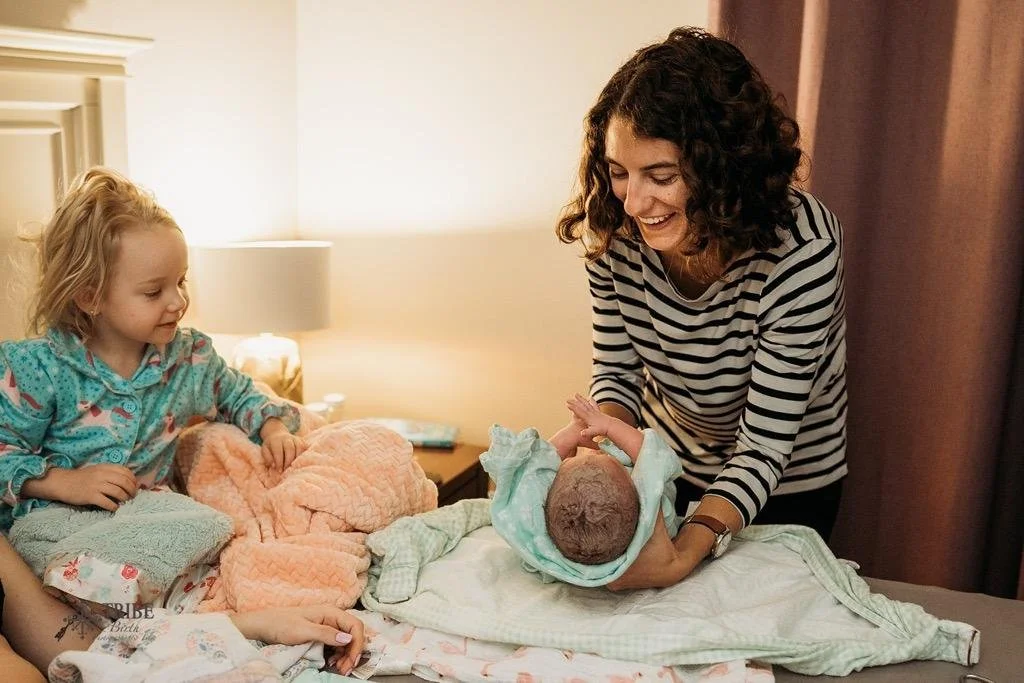 A woman with curly hair holding a newborn baby, with a young girl sitting nearby, in a cozy bedroom setting.