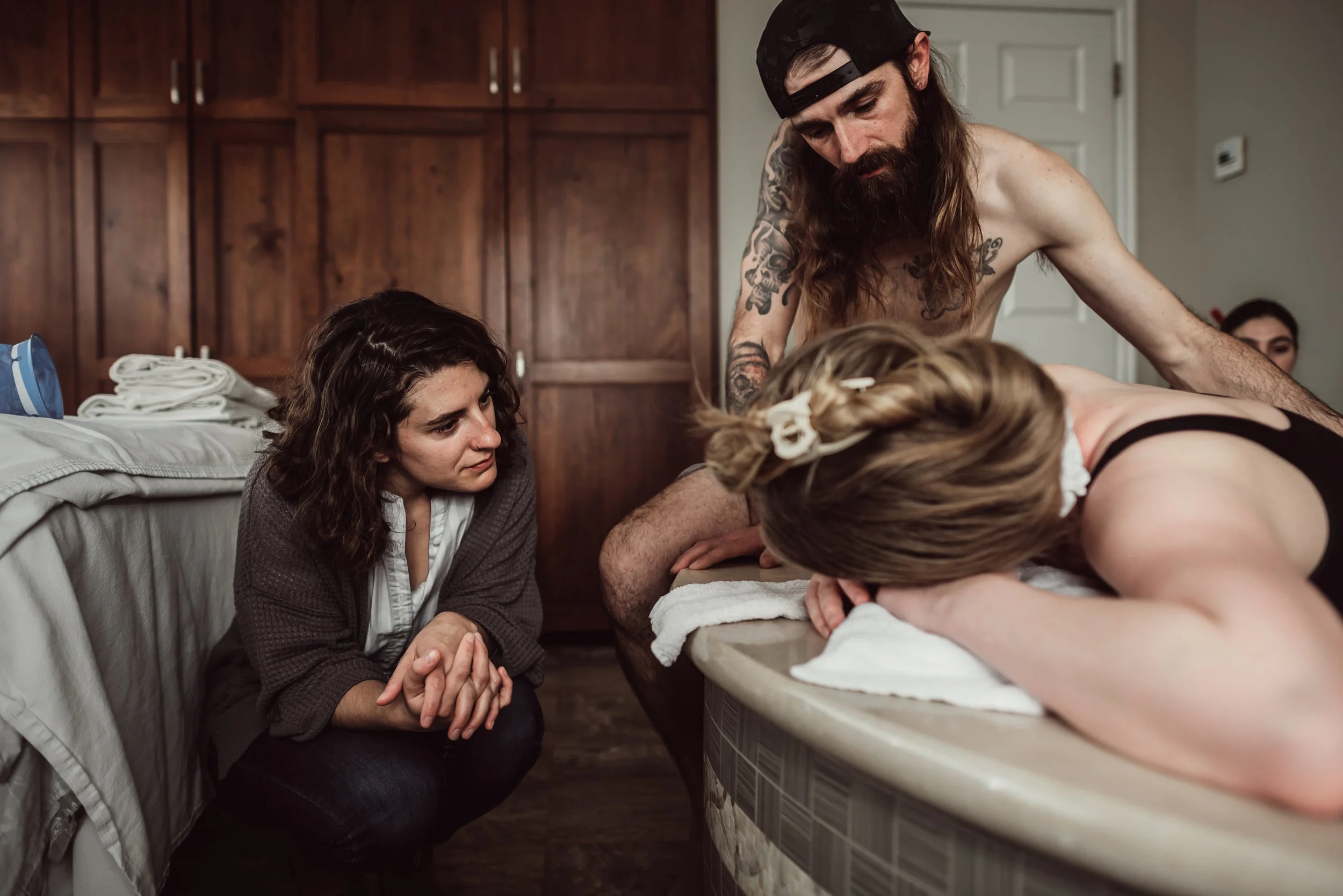 Group of people in a room, with women kneeling and praying, while a shirtless man with long hair and tattoos leans over a woman lying on a table.