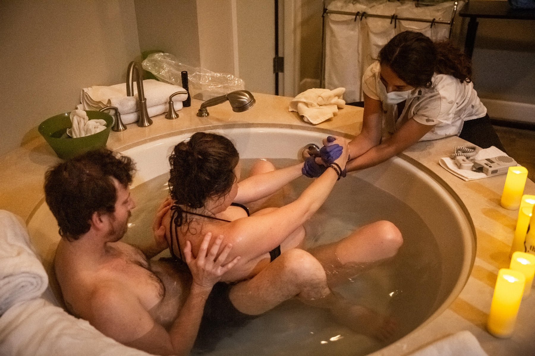 People having a bath together in a large bathtub with candles around the edge, with one person helping another in the tub.