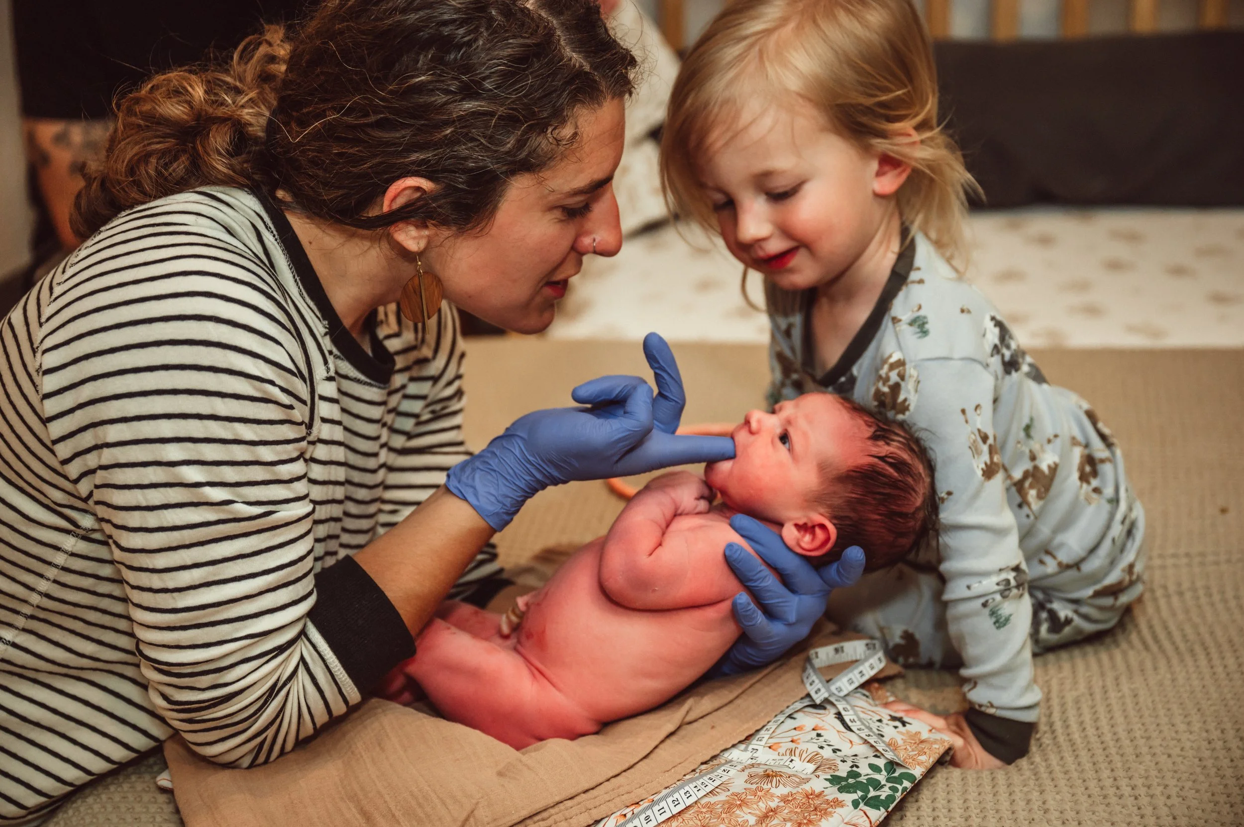 Woman, wearing a striped shirt and gloves, examining a newborn baby with a little girl, in a cozy indoor setting with a bed and measuring tape in the background.