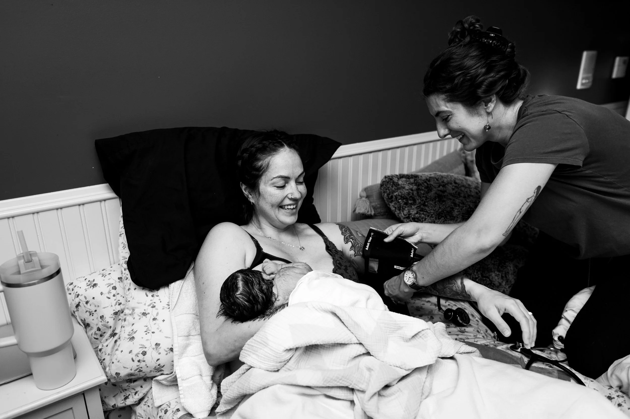 A woman with tattoos on her arm is lying in bed, smiling, as a nurse or caregiver with short hair and earrings interacts with her, possibly during the birth of a baby.