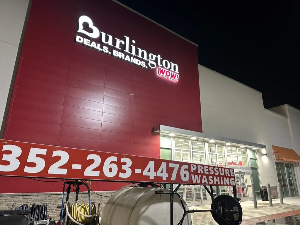 Exterior of Burlington store at night with signage for deals, brands, and a WOW promotion, with pressure washing equipment and a large red banner displaying a phone number.