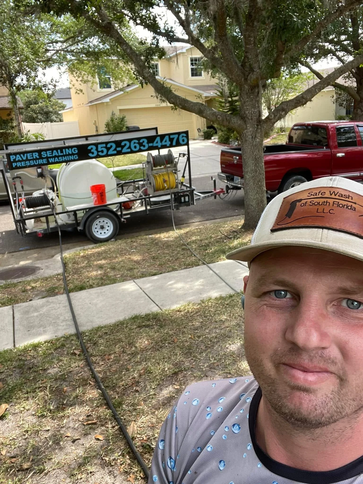 A man taking a selfie outdoors on a sidewalk with a paver sealing trailer and a red truck in the background, under a large tree in a residential neighborhood.