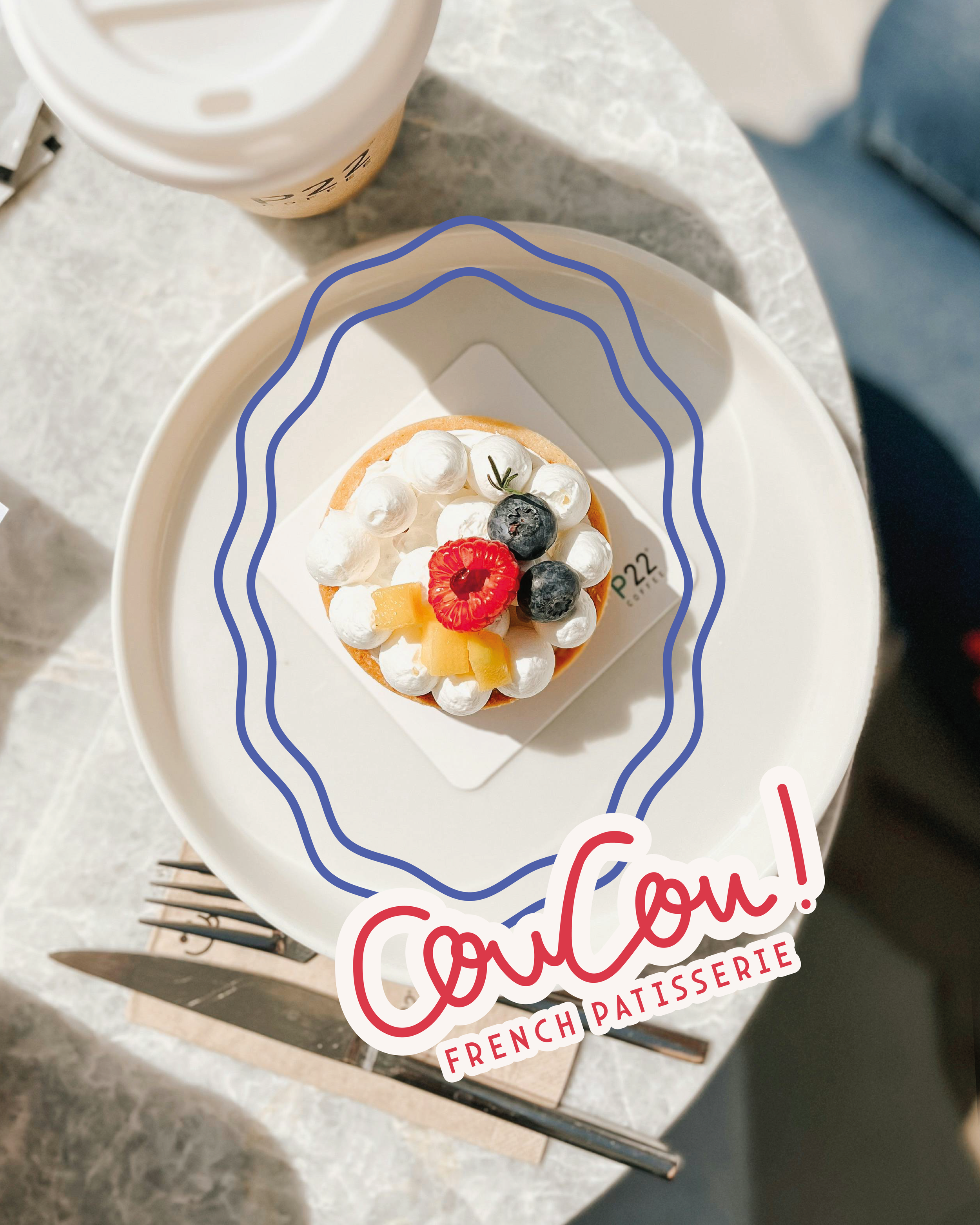 A fruit tart topped with whipped cream, blueberries, raspberry, pineapple, and a sprig of rosemary, placed on a white plate with a sticker that says 'Coucou! French Patisserie.'
