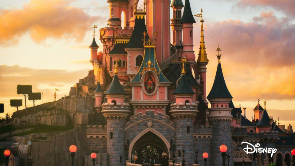 Disney castle with pink and blue turrets at sunset, surrounded by trees and lit lanterns.