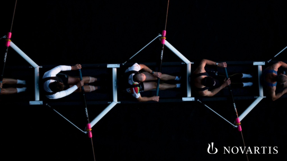 Four rowers in a racing shell, viewed from above, navigating a dark water course during a competitive rowing event.