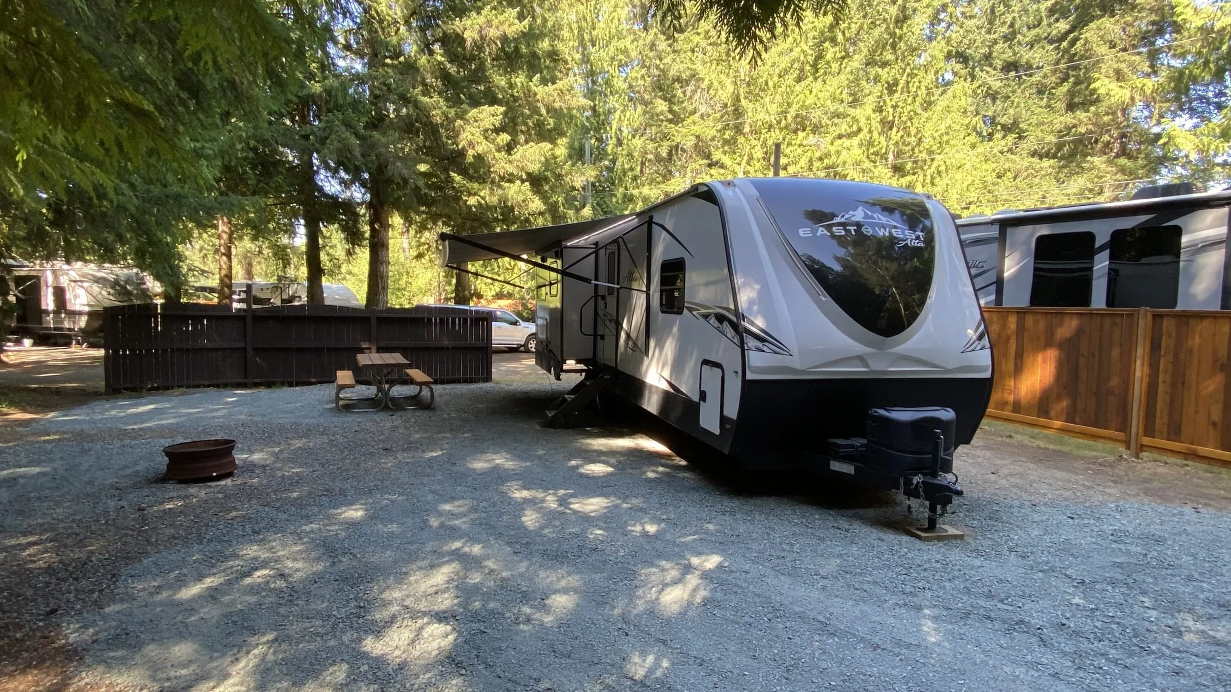 A travel trailer parked in a wooded campsite with trees and a wooden fence in the background. There is a picnic table and a fire ring in the gravel area.