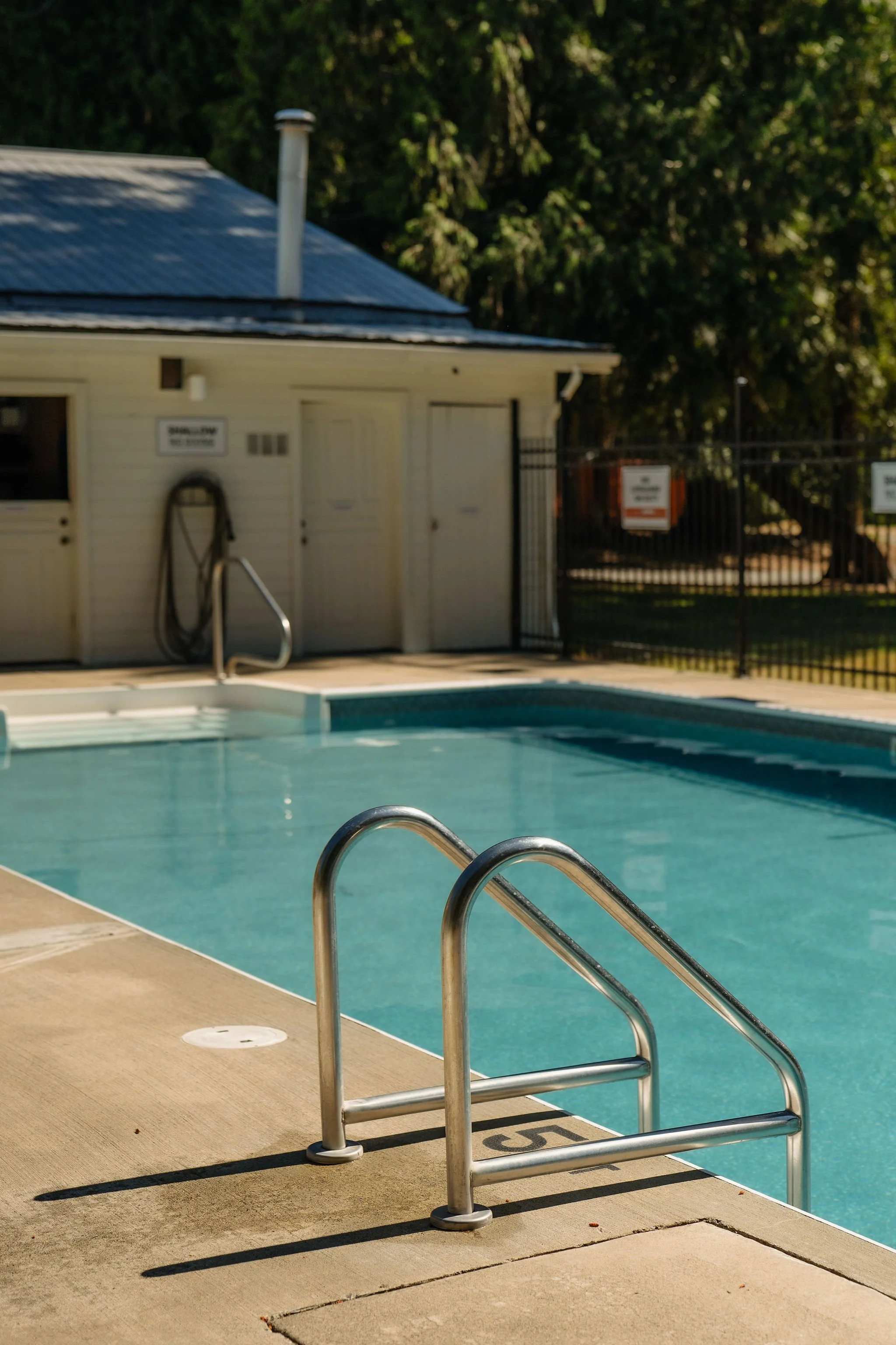 An outdoor swimming pool with two people relaxing in it. Chairs and umbrellas are around the pool, with a building behind, set against a backdrop of wooded trees and a clear blue sky.
