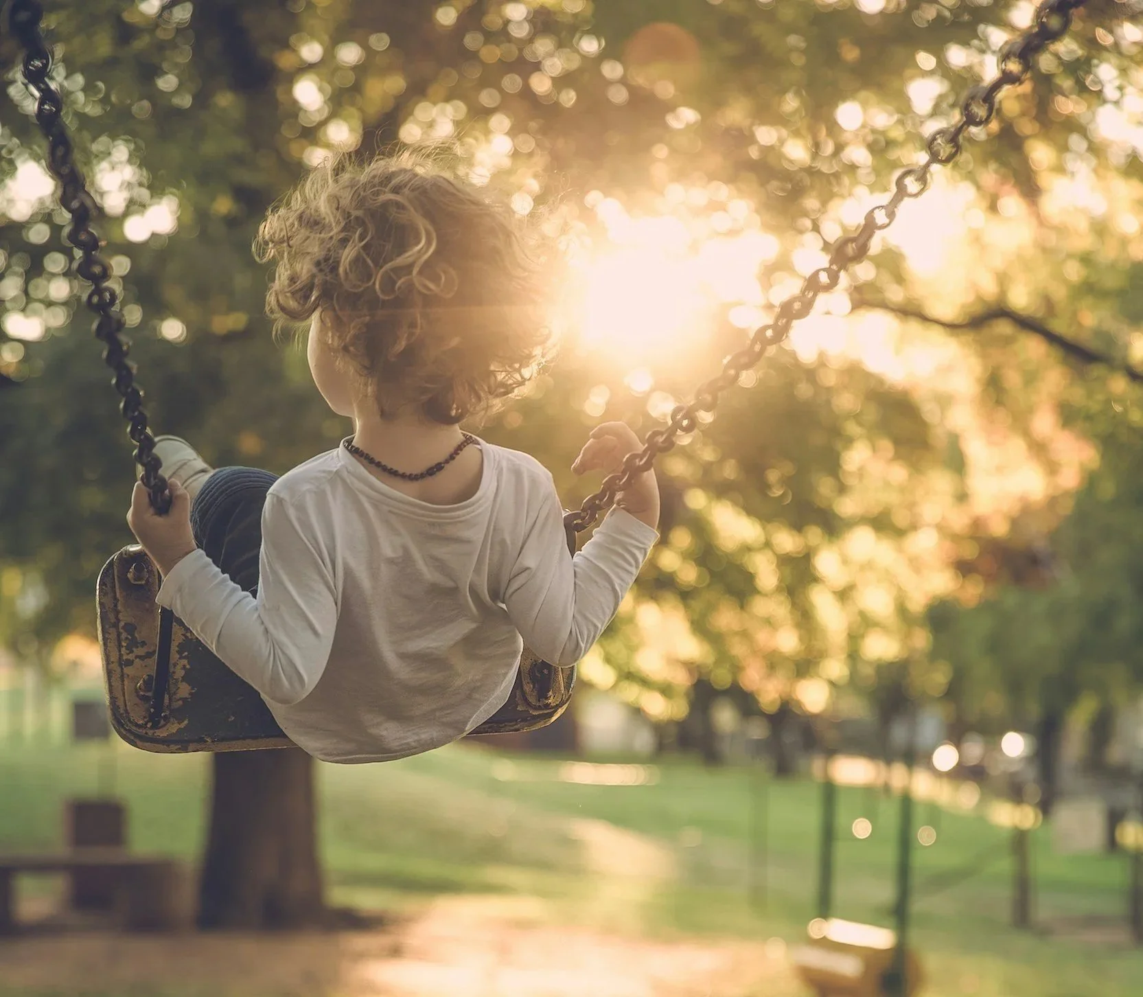 Child on swing in park playground with sunlight streaming through trees