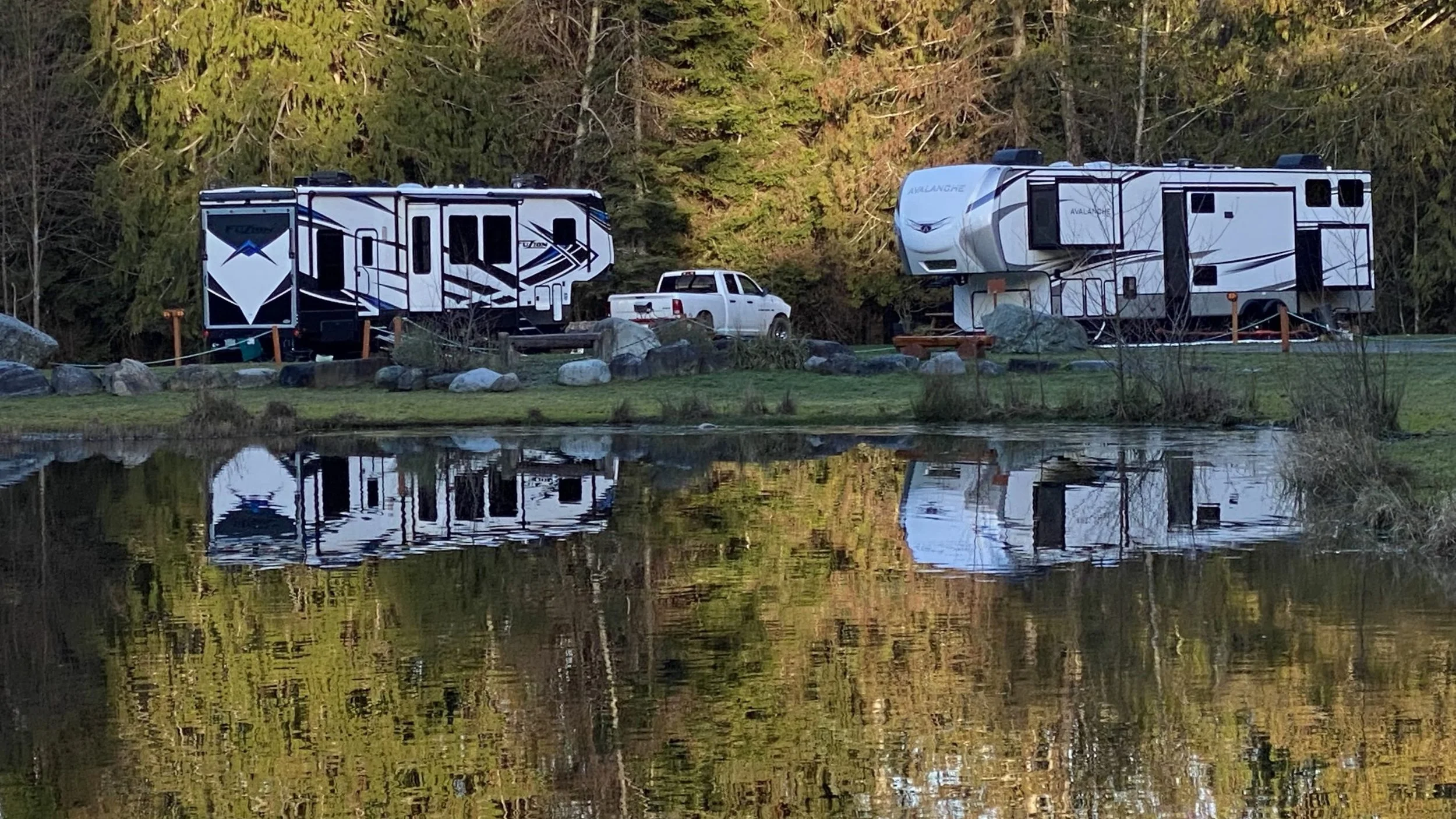 Two large RVs parked near a pond with trees in the background, and a white pickup truck between them.