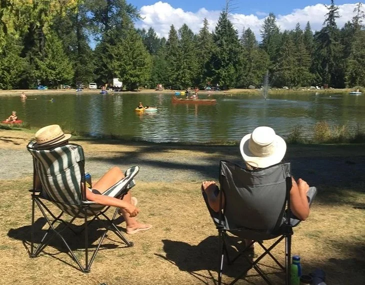 Two people sitting in camping chairs by a lake, wearing wide-brimmed hats, with trees and people in boats and swimming in the background.