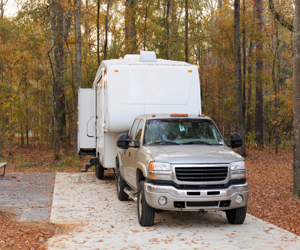 A beige pickup truck with a large white RV attached, parked on a gravel and concrete pathway in a wooded area with autumn foliage.