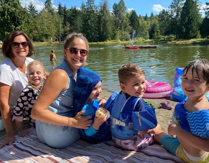 A family enjoying a day at the lake, sitting on a blanket near the water with children in and out of the lake, surrounded by trees and a boat in the background.