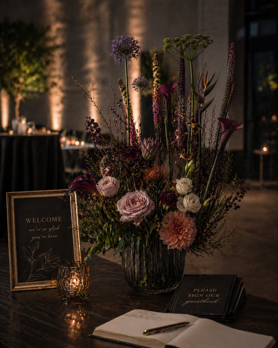 A wedding reception registration table with a large floral arrangement of red roses, purple hydrangeas, and dark purple flowers, surrounded by candles, a guest sign-in book, and signs for registration and gifts.