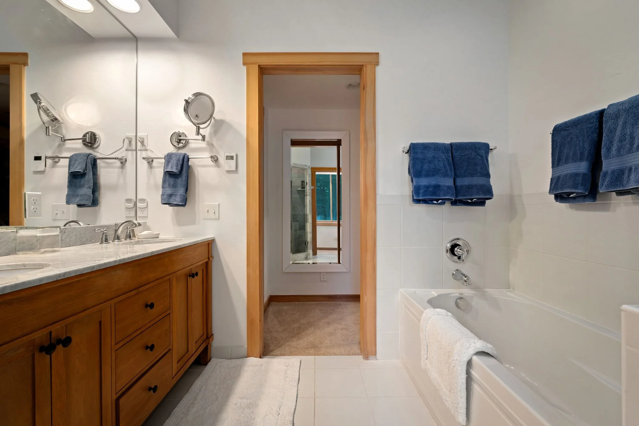 A bathroom with a wooden vanity, a marble countertop, and a large mirror. There are blue towels hanging on the wall and on towel racks. A white bathtub with a white towel folded over the side. A doorway leads to another room with a mirror reflecting 