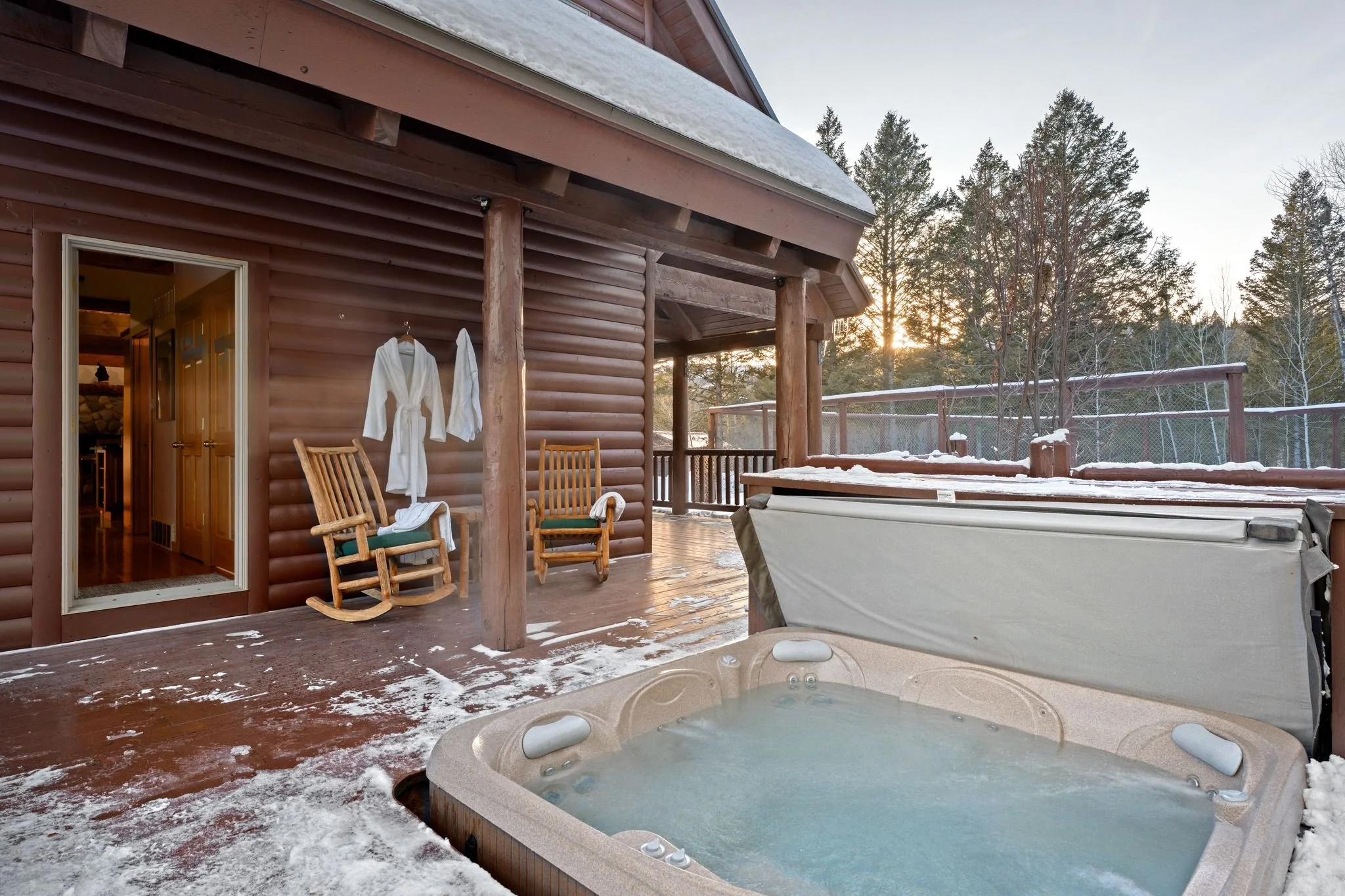 A snowy outdoor hot tub on a wooden deck with two wooden chairs, bathrobes hanging on the wall, and a view of trees at sunset.