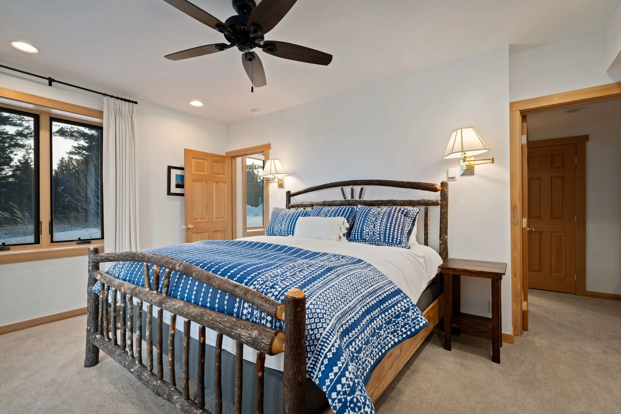 Bedroom with a rustic wooden bed, blue and white patterned bedding, twin side tables, wall-mounted lamps, window with white curtains, ceiling fan, and wooden door.