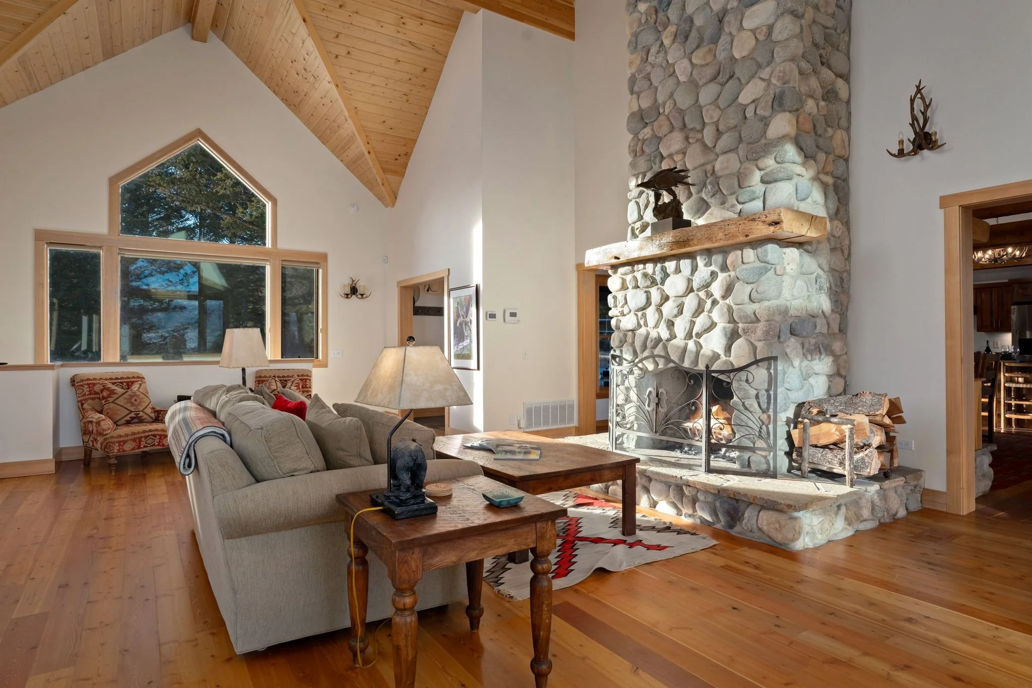 Living room featuring a large stone fireplace, beige sofa, wooden side tables, and a patterned area rug. Large windows with wooden trim and a vaulted wooden ceiling enhance the space.