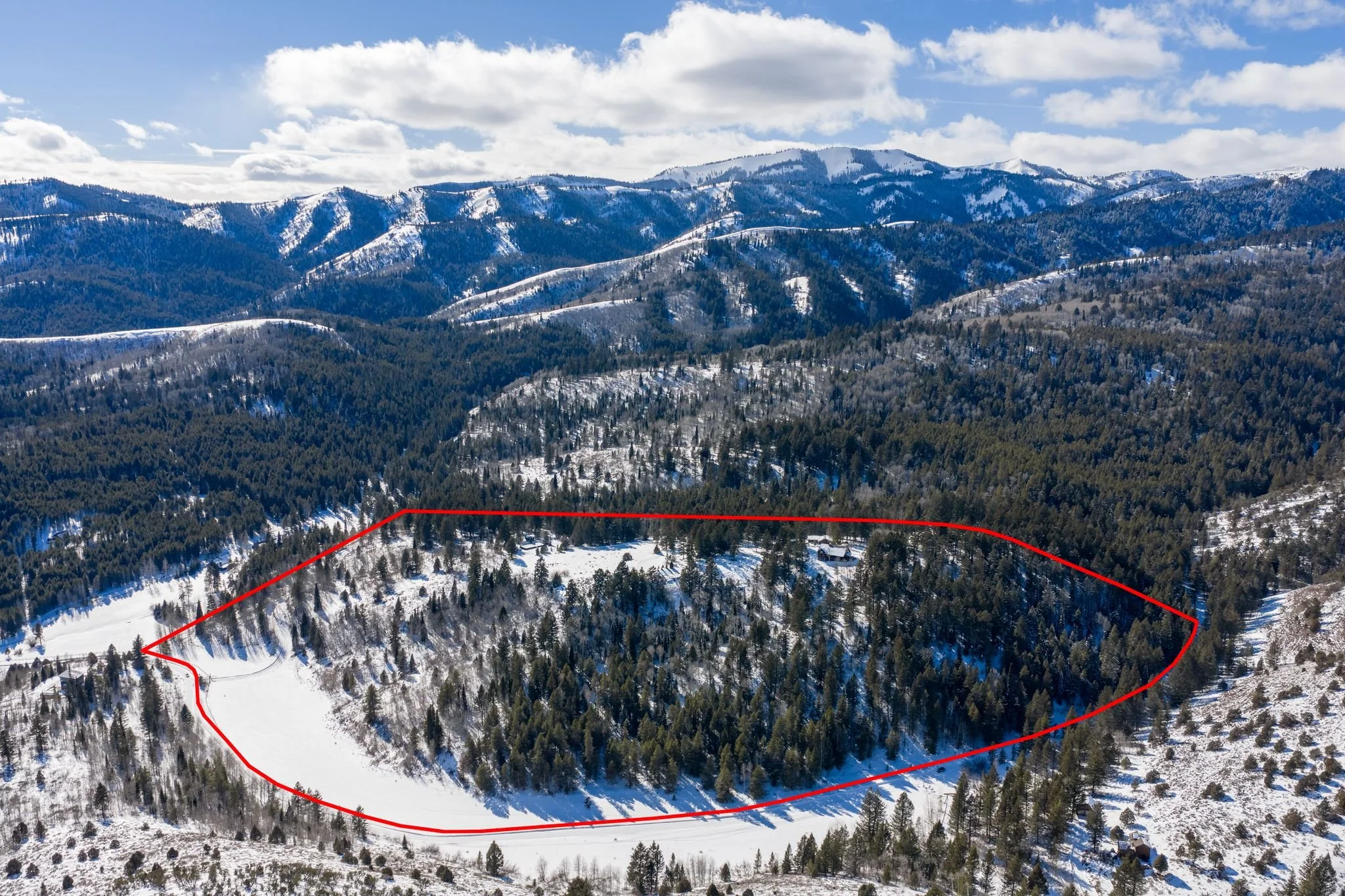 Snow-covered mountain landscape with a forested area outlined in red on a sunny day with clouds in the sky.