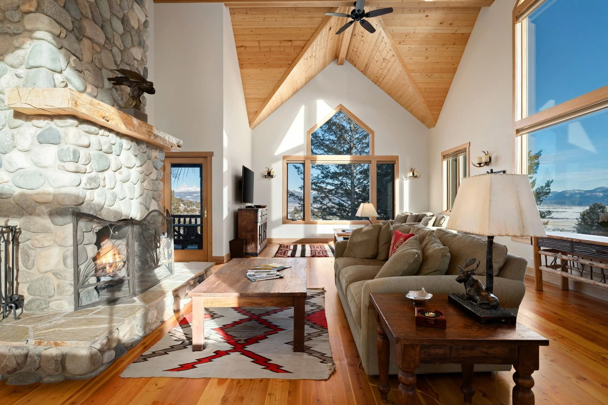 Living room with stone fireplace, wooden ceiling, large windows with mountain view, beige sofa, wooden coffee table, patterned rug, TV stand, and decorative items.