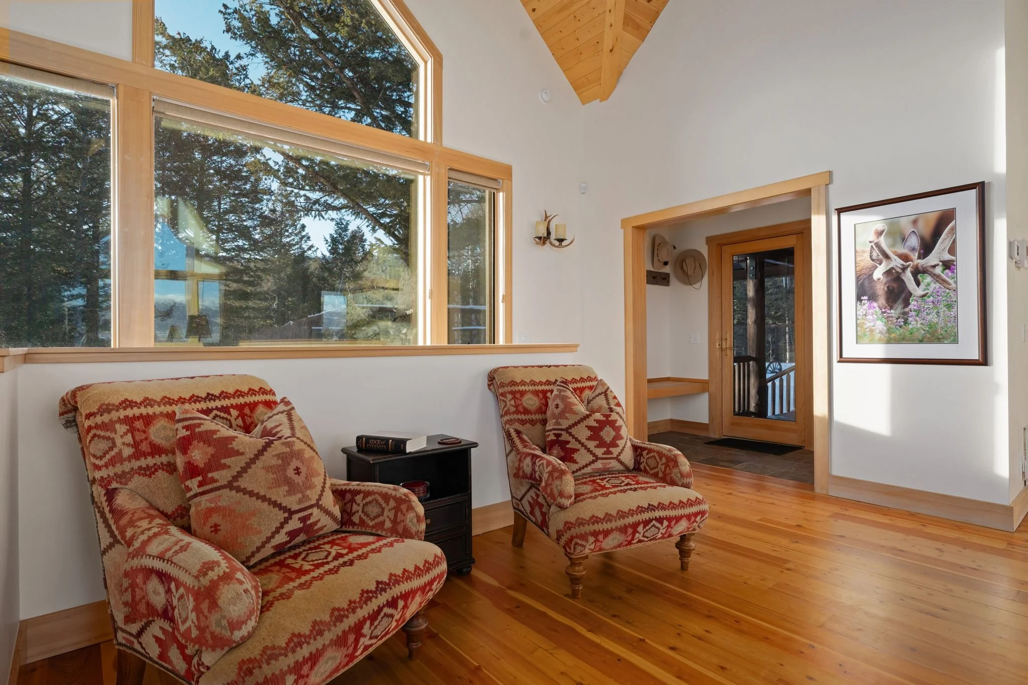 Living room with large windows showing trees outside, two patterned armchairs, a small black side table with books, and a framed moose and deer artwork on the wall.