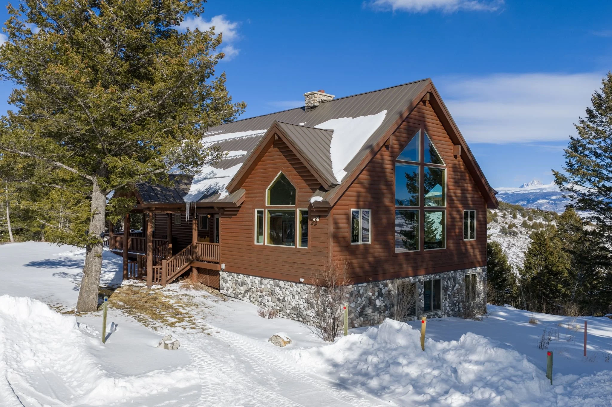 A wooden house with large windows, surrounded by snow-covered ground and trees, with a mountain range in the background under a blue sky.